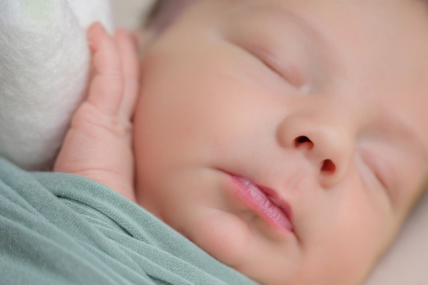 Peaceful newborn baby sleeping soundly, wrapped in a soft green blanket with tiny hand raised near face.
