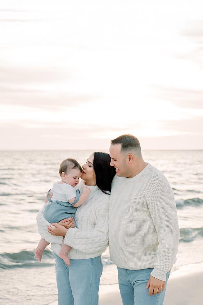 Family of three poses on beach at sunset, parents in white sweaters and jeans, mother kissing baby.