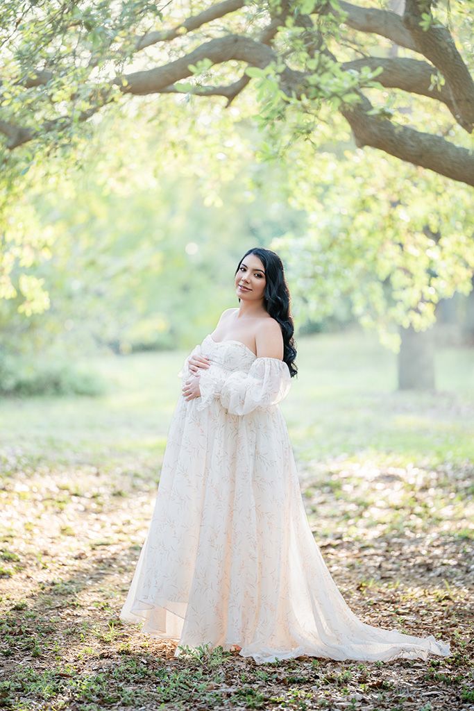 Pregnant woman in elegant white lace off-shoulder gown posing outdoors under green trees in soft natural light.