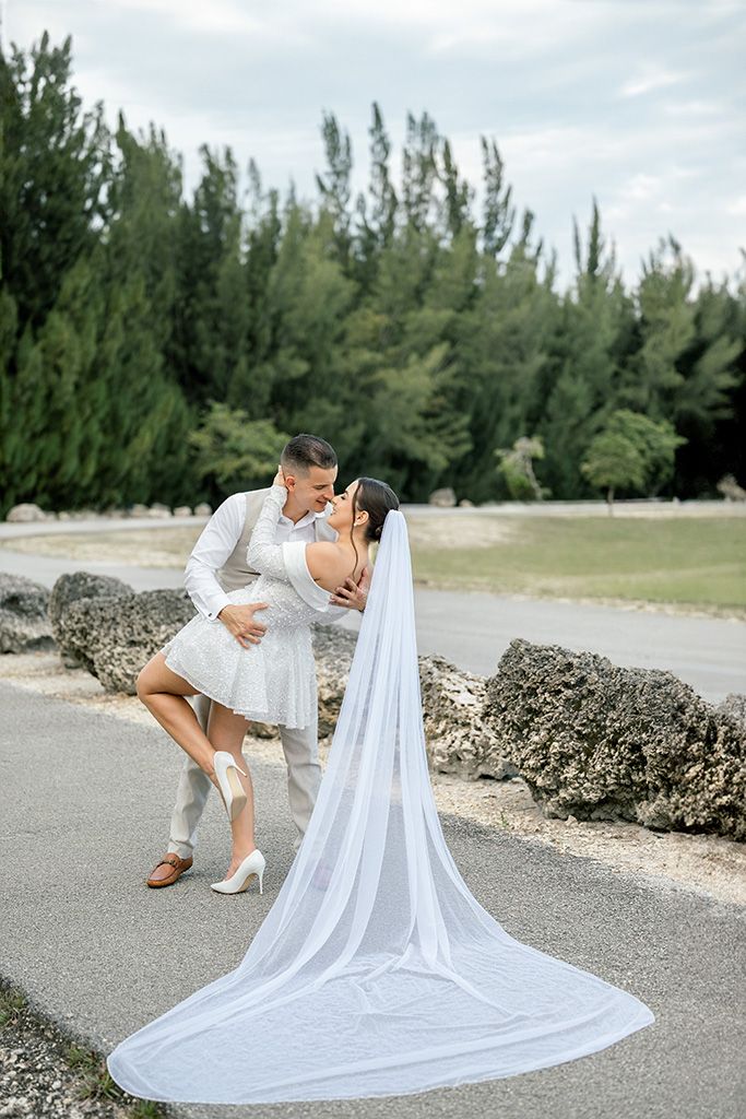 Groom dipping bride in a romantic kiss, bride in short white dress with long veil, outdoor wedding photo.