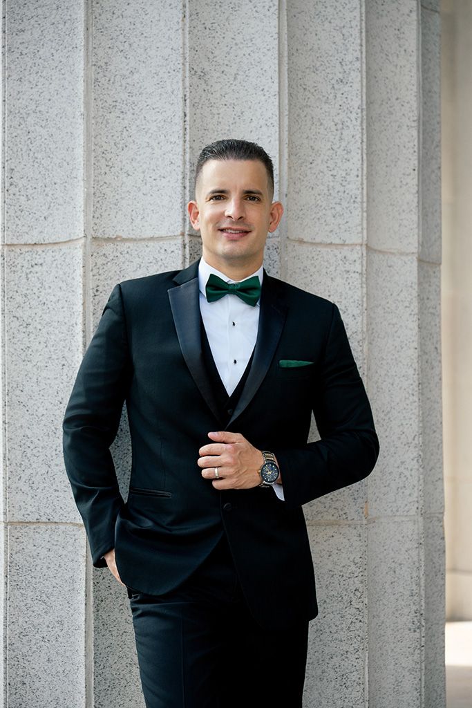 Smiling groom in black tuxedo with green bow tie and pocket square poses by stone columns outdoors.