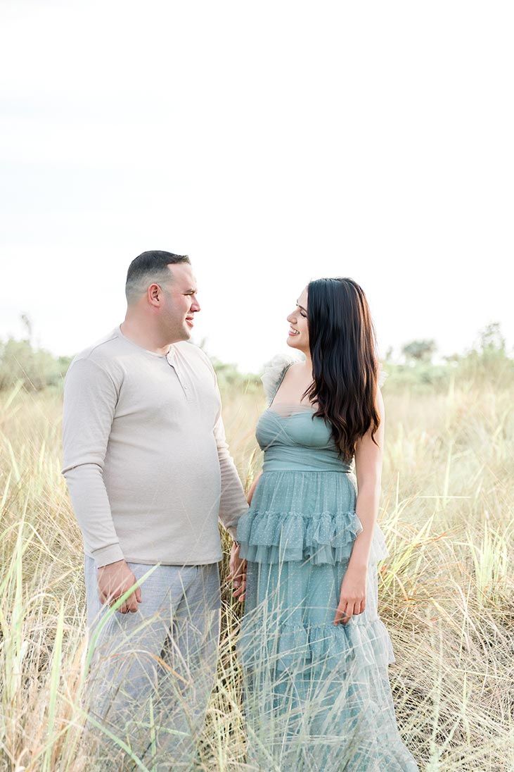Couple holding hands and smiling at each other in tall golden grass field, woman in teal dress, outdoor portrait.