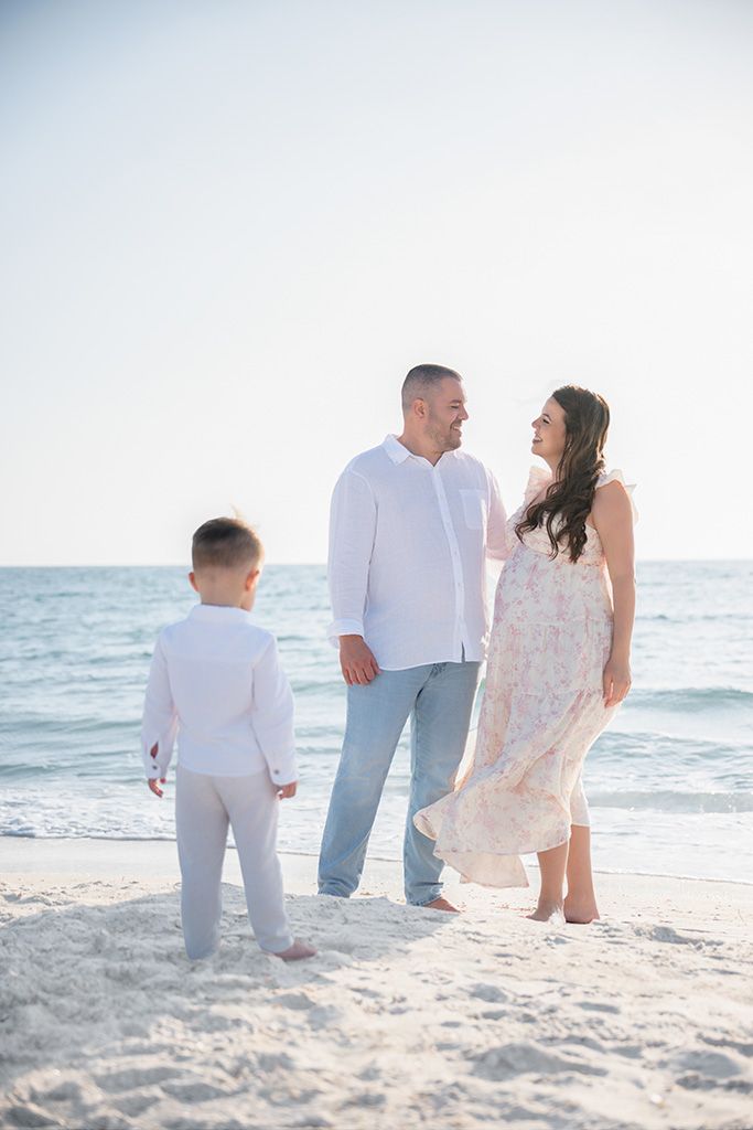 Family of three in white outfits enjoying a sunny beach day, parents smiling at each other while child stands nearby.
