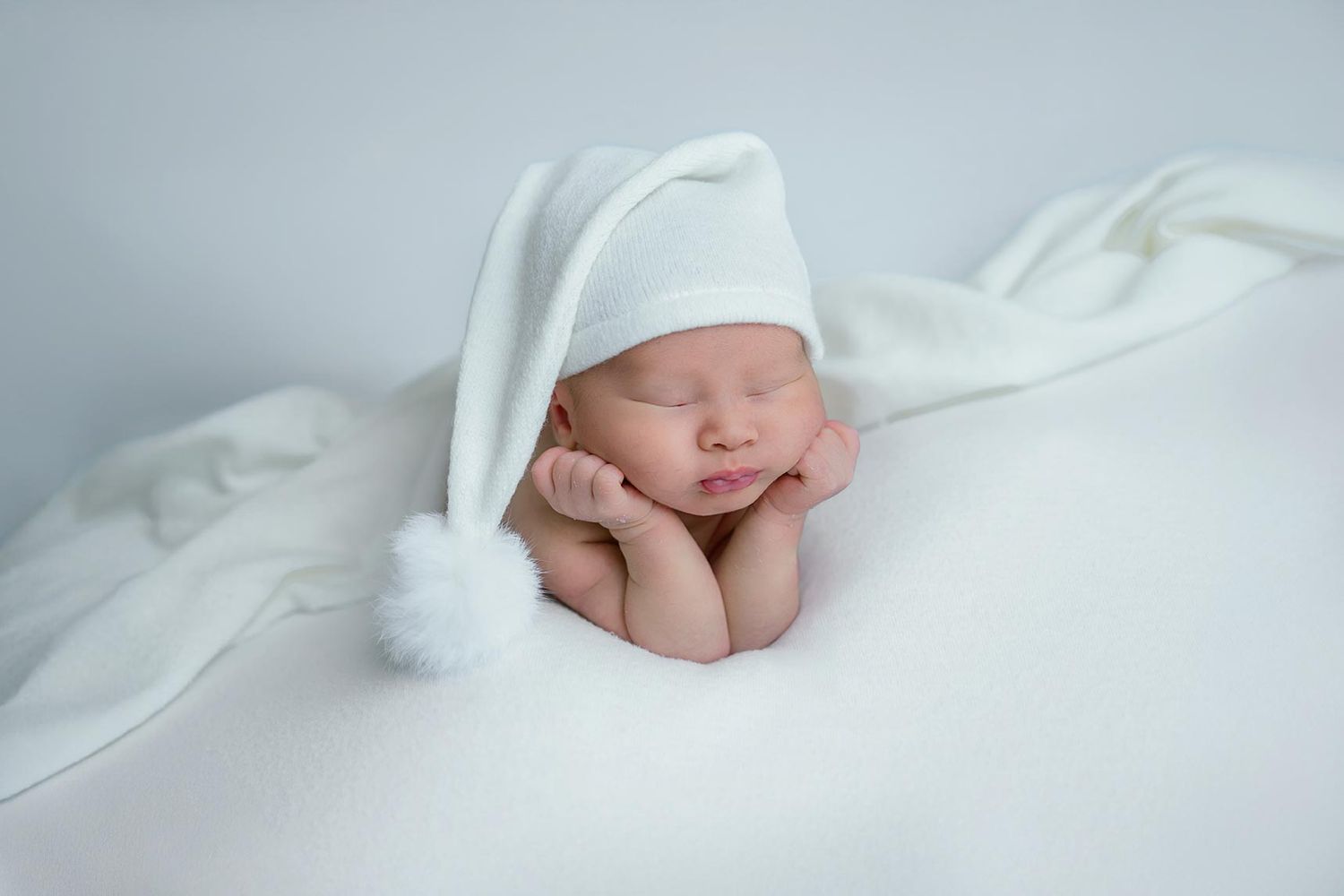 Newborn baby sleeping peacefully on white fabric wearing a white pom pom hat.