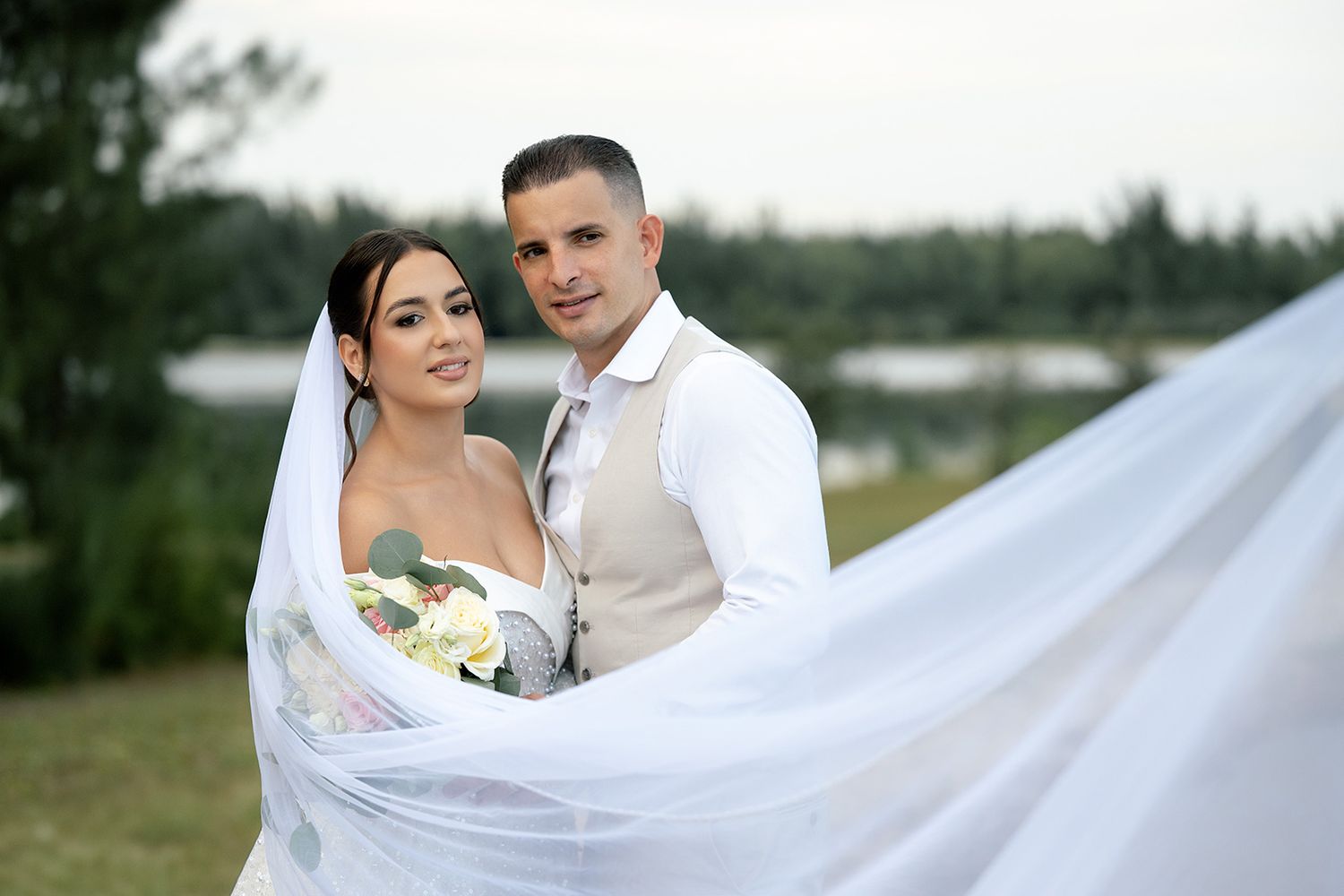 Bride and groom pose outdoors near a lake, her flowing white veil billowing in the wind on their wedding day.