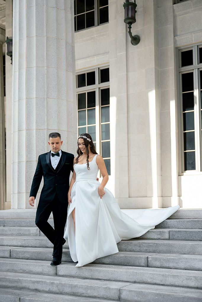 Bride and groom descend stone steps of elegant building, she in flowing white gown, he in classic black tuxedo.