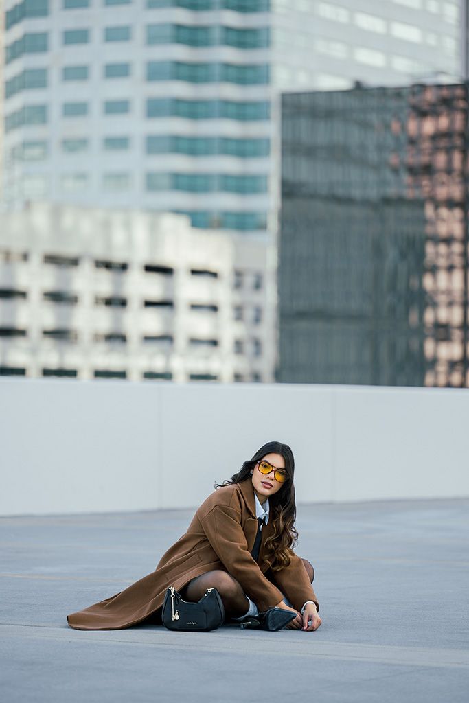 Woman in brown coat and yellow sunglasses sitting on rooftop with city buildings in background.