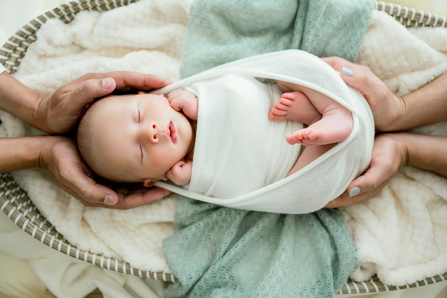 Gentle hands cradle a peaceful sleeping newborn baby wrapped in soft white swaddling cloth.