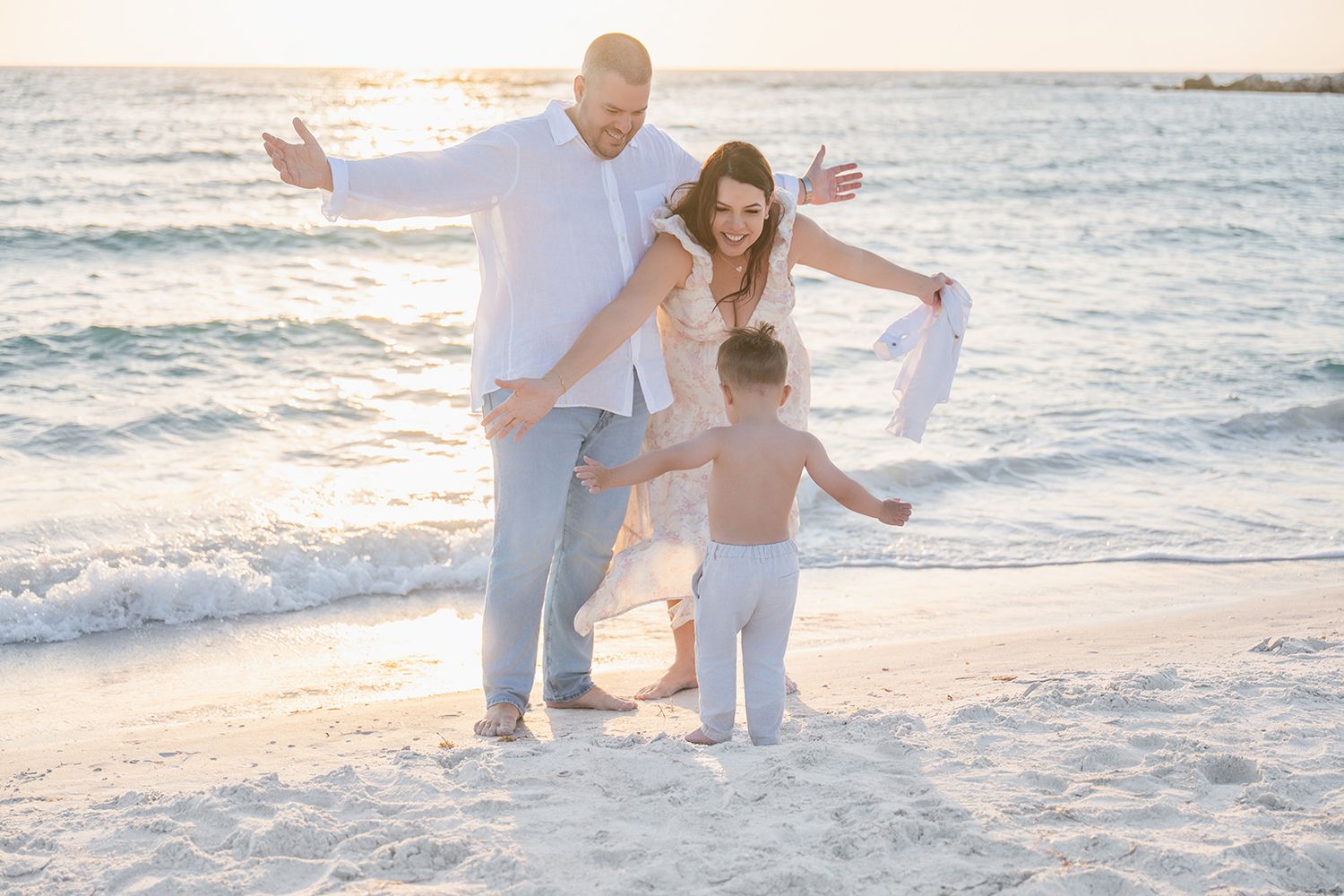 Joyful family with toddler celebrating on sunny Florida beach with turquoise ocean waves behind them.