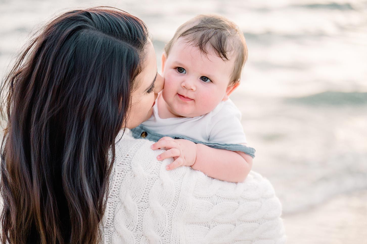 A mother in a white sweater kisses her baby on the cheek at the beach.