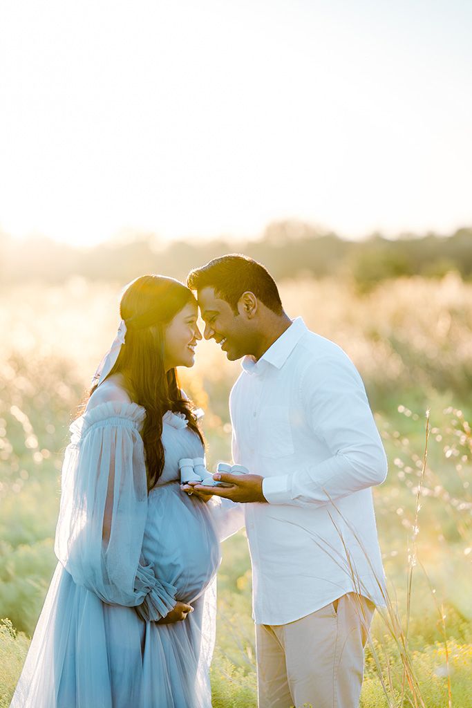 Pregnant couple sharing a tender moment in a sunlit field, woman in blue dress holding baby shoes.