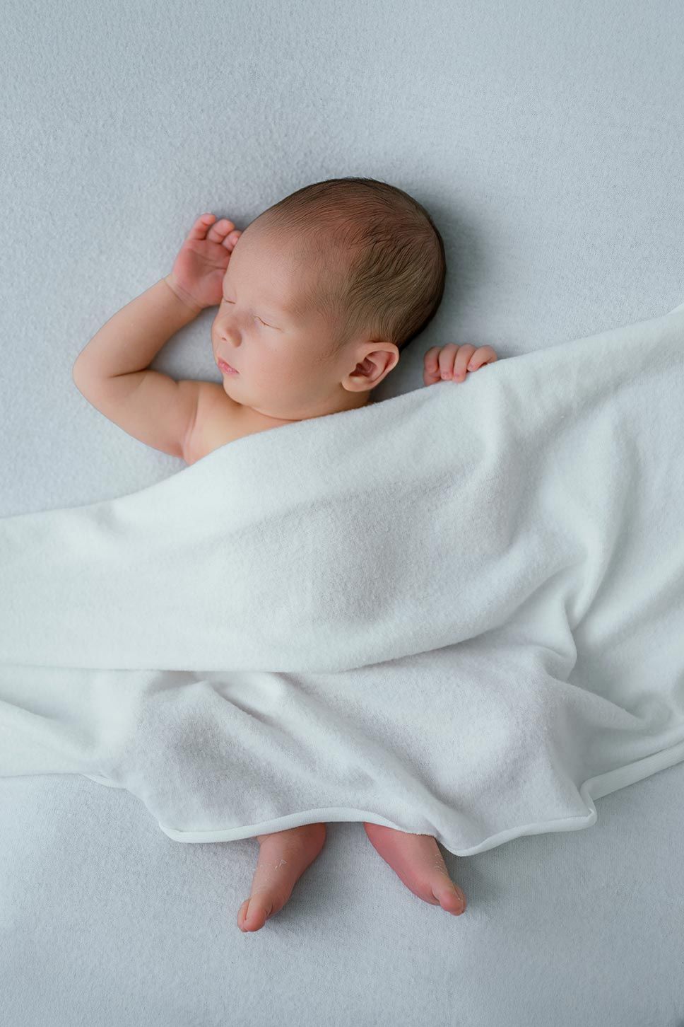Peaceful newborn baby sleeping on a light blue surface, wrapped in a soft white blanket with tiny feet peeking out.