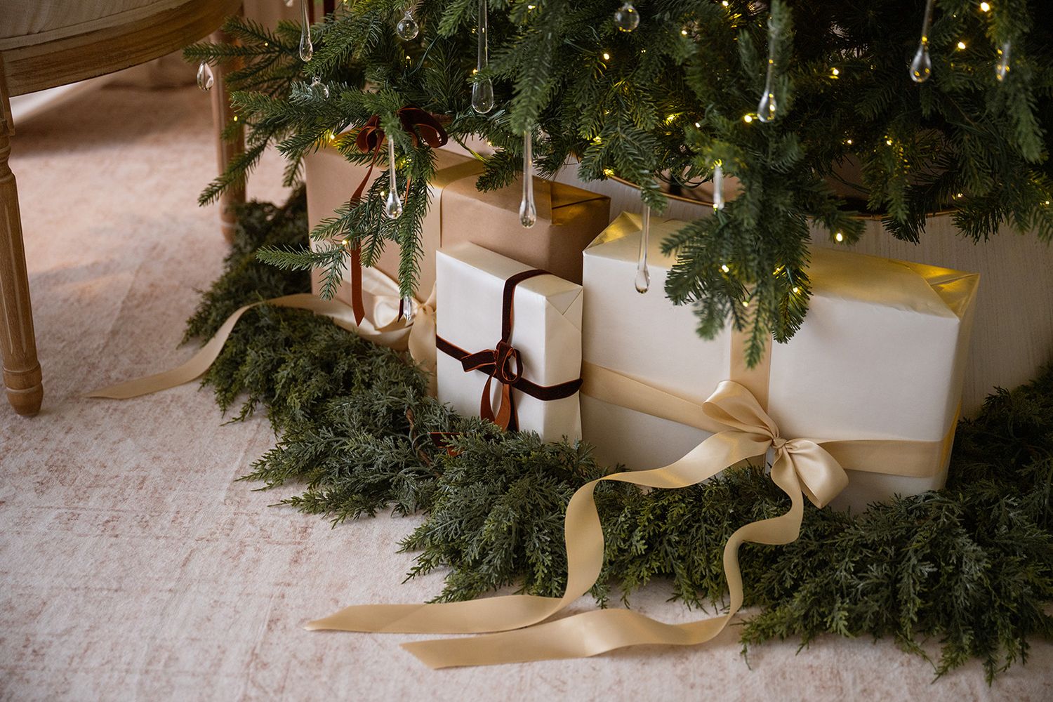 Elegant white Christmas gifts with gold and burgundy ribbons arranged under a decorated pine tree with garland.
