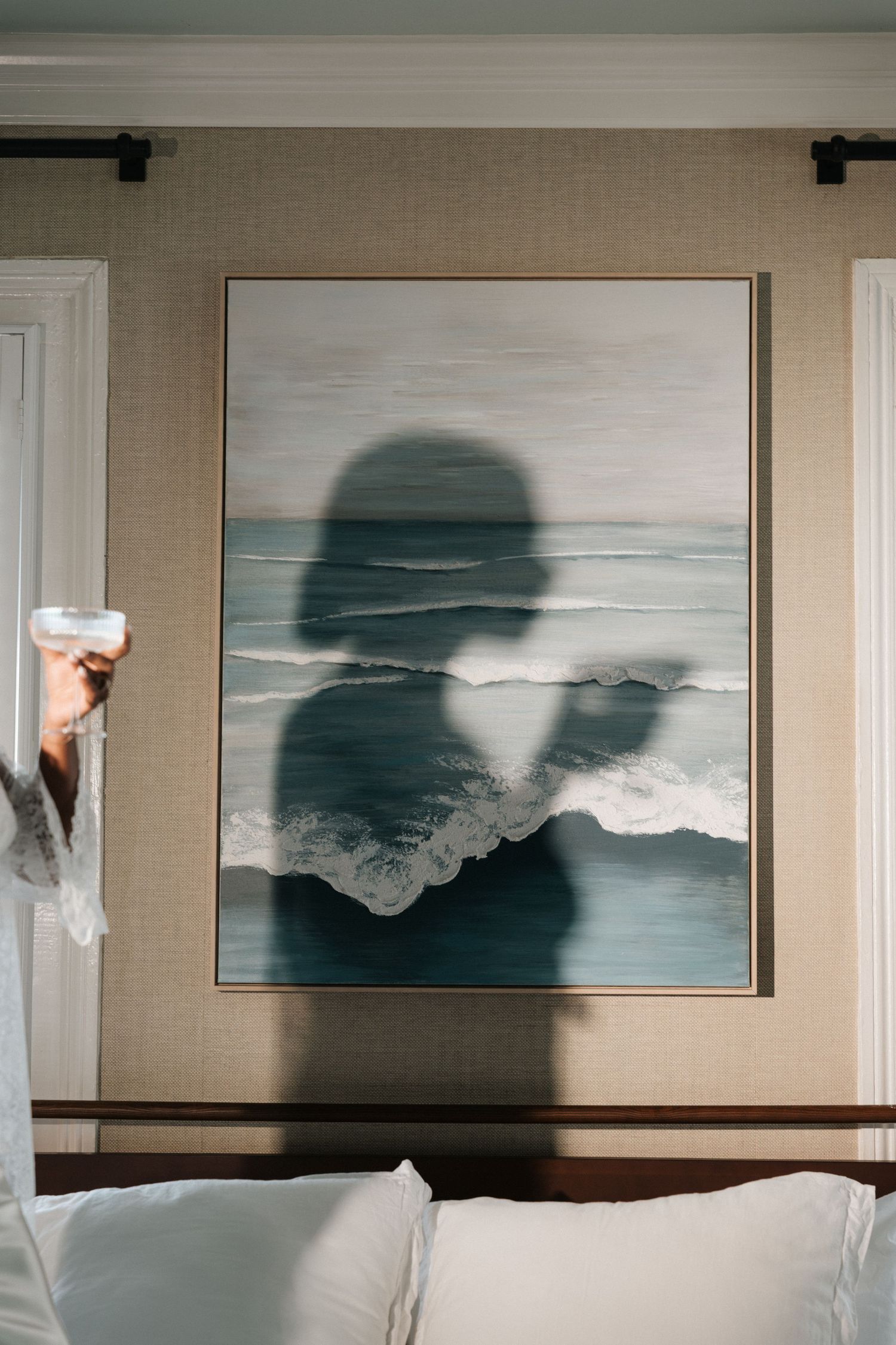 Shadow of a person silhouetted against a large ocean wave painting hanging above a bed in a neutral bedroom.