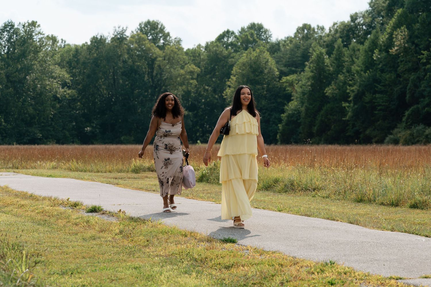 Two women in maxi dresses walk hand-in-hand on a path through a scenic field with lush green trees.