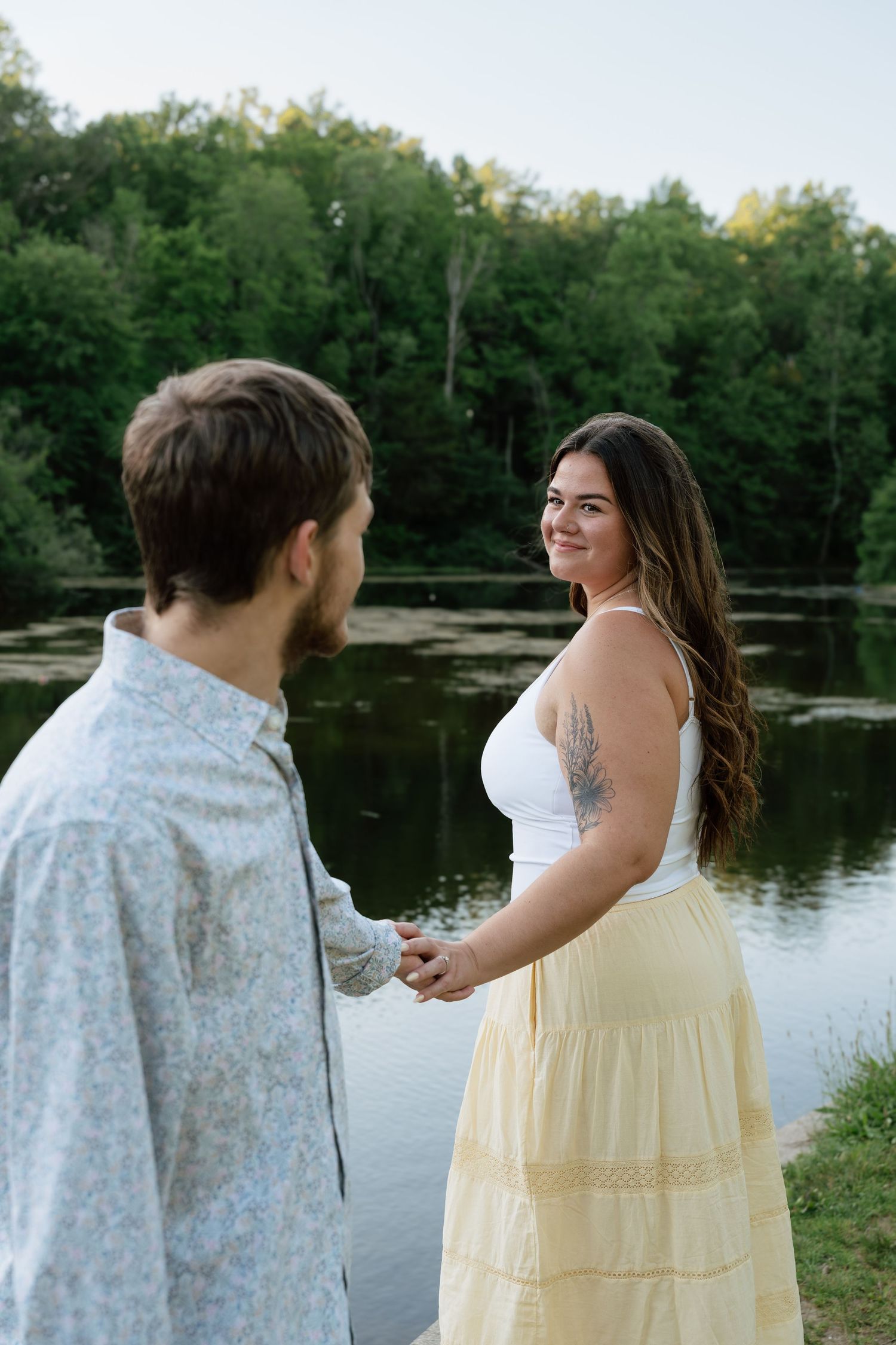 Smiling brunette woman in white top and yellow skirt holds hands with man by a serene lake with green trees.