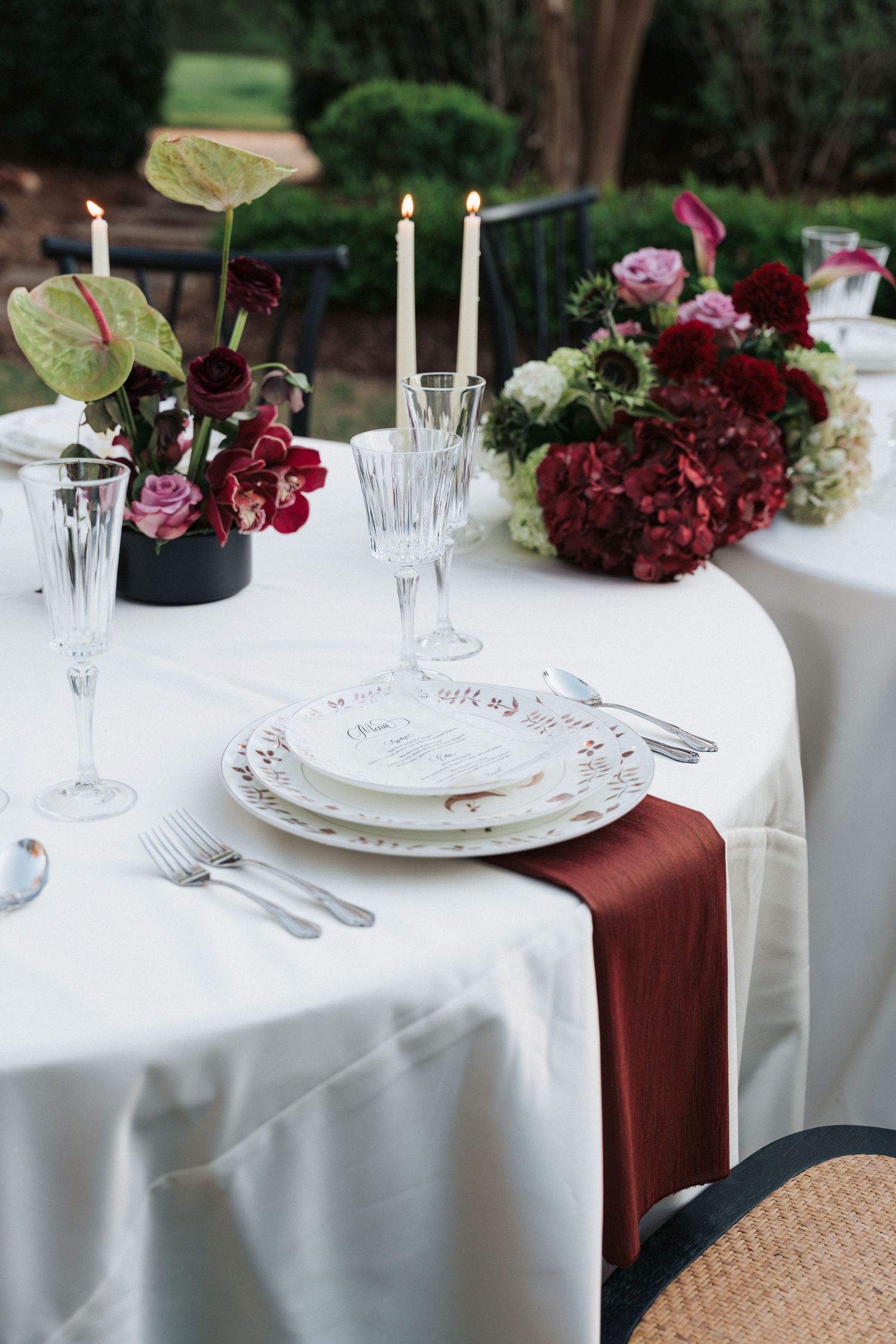 Elegant wedding table setting with burgundy napkins, crystal glasses, white plates, and floral centerpieces of red hydrangeas and roses.