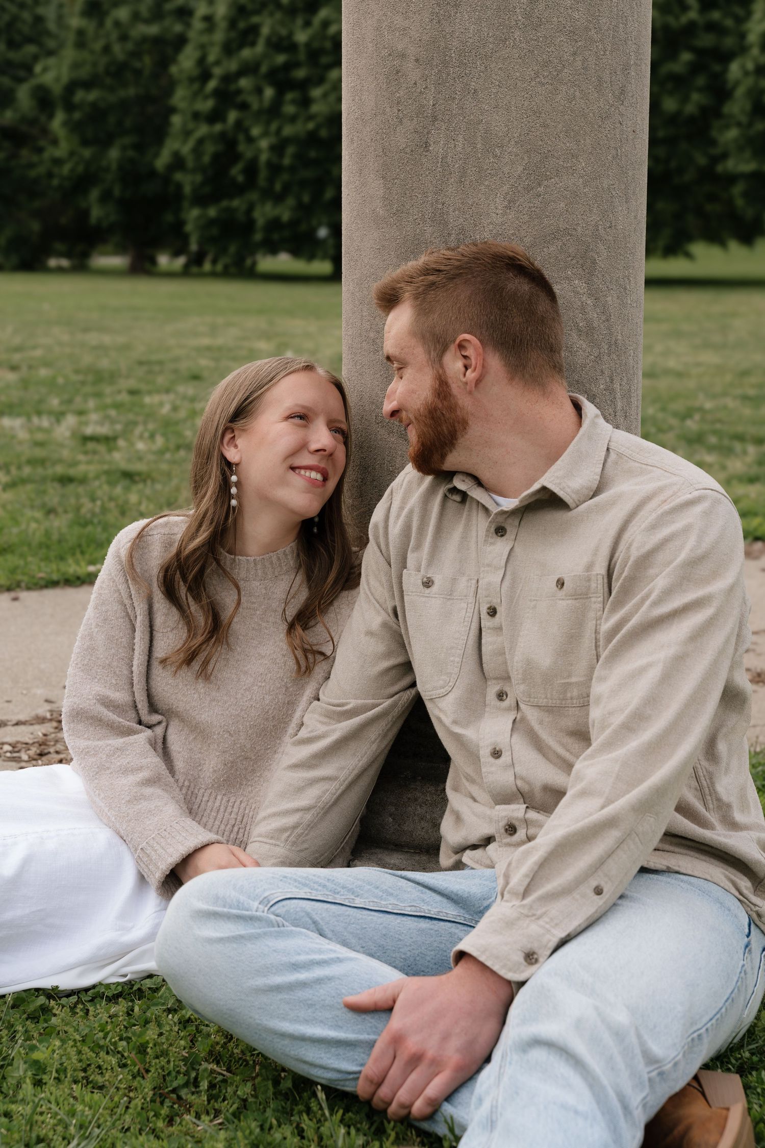 A smiling couple sits together against a concrete pillar in a park, dressed in neutral tones, sharing a warm moment.
