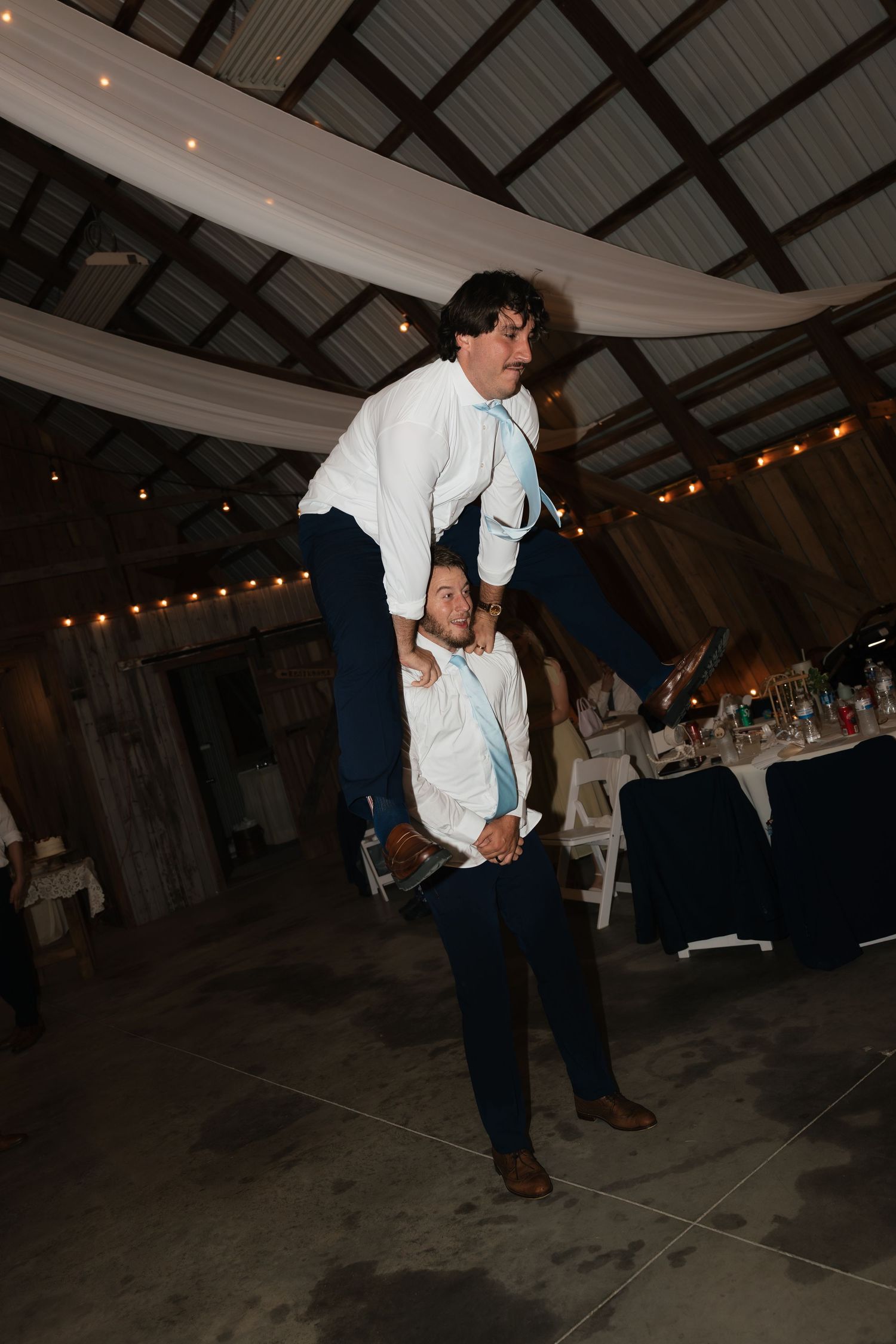 Wedding guest performs impressive shoulder lift stunt on dance floor inside rustic barn reception venue.