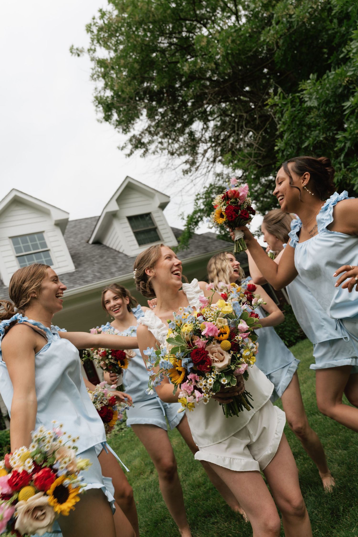 Bridesmaids in light blue outfits laugh and celebrate outdoors holding colorful wildflower bouquets with a bride in white.