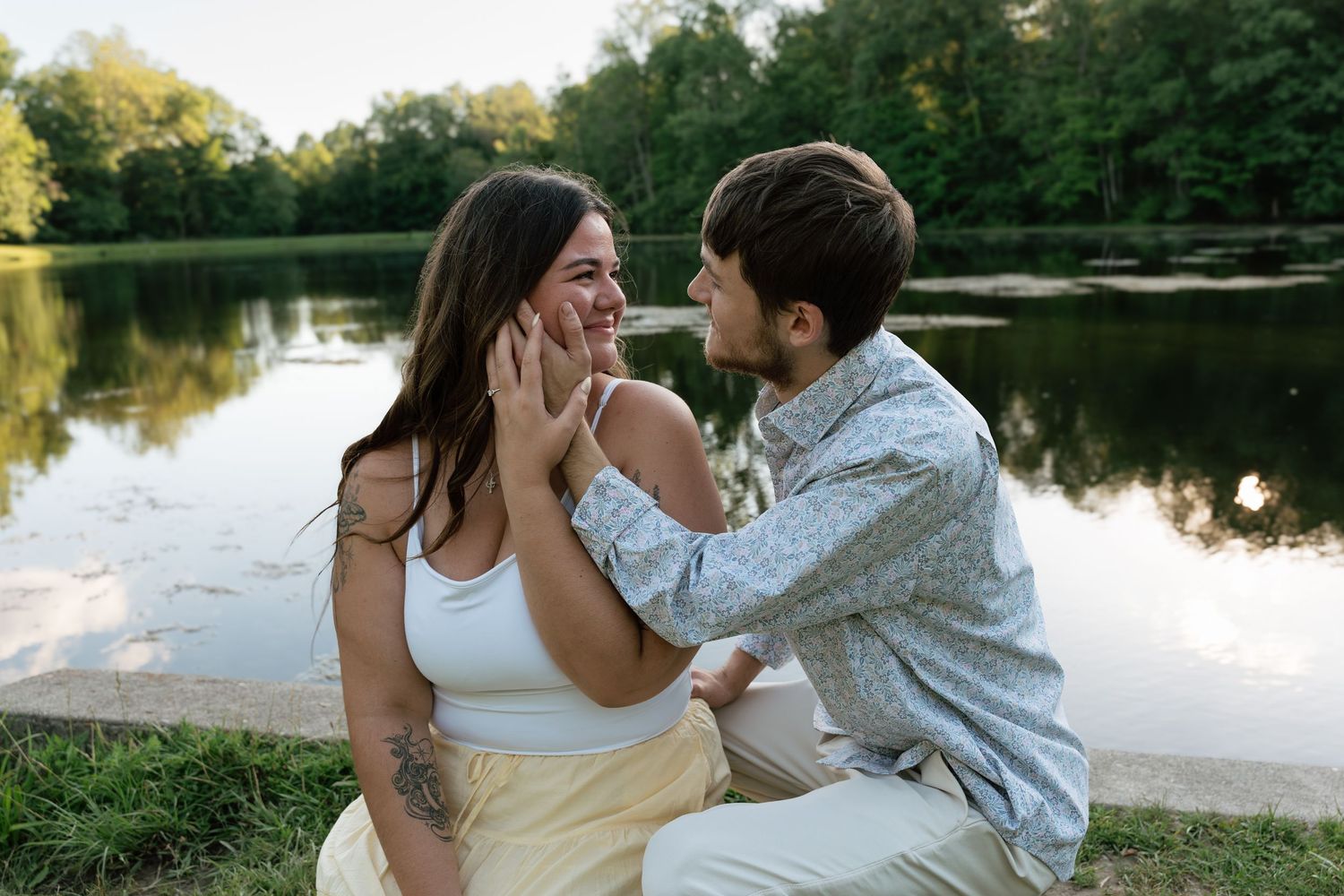 Couple sharing a romantic moment by a serene lake, surrounded by lush green trees at dusk.
