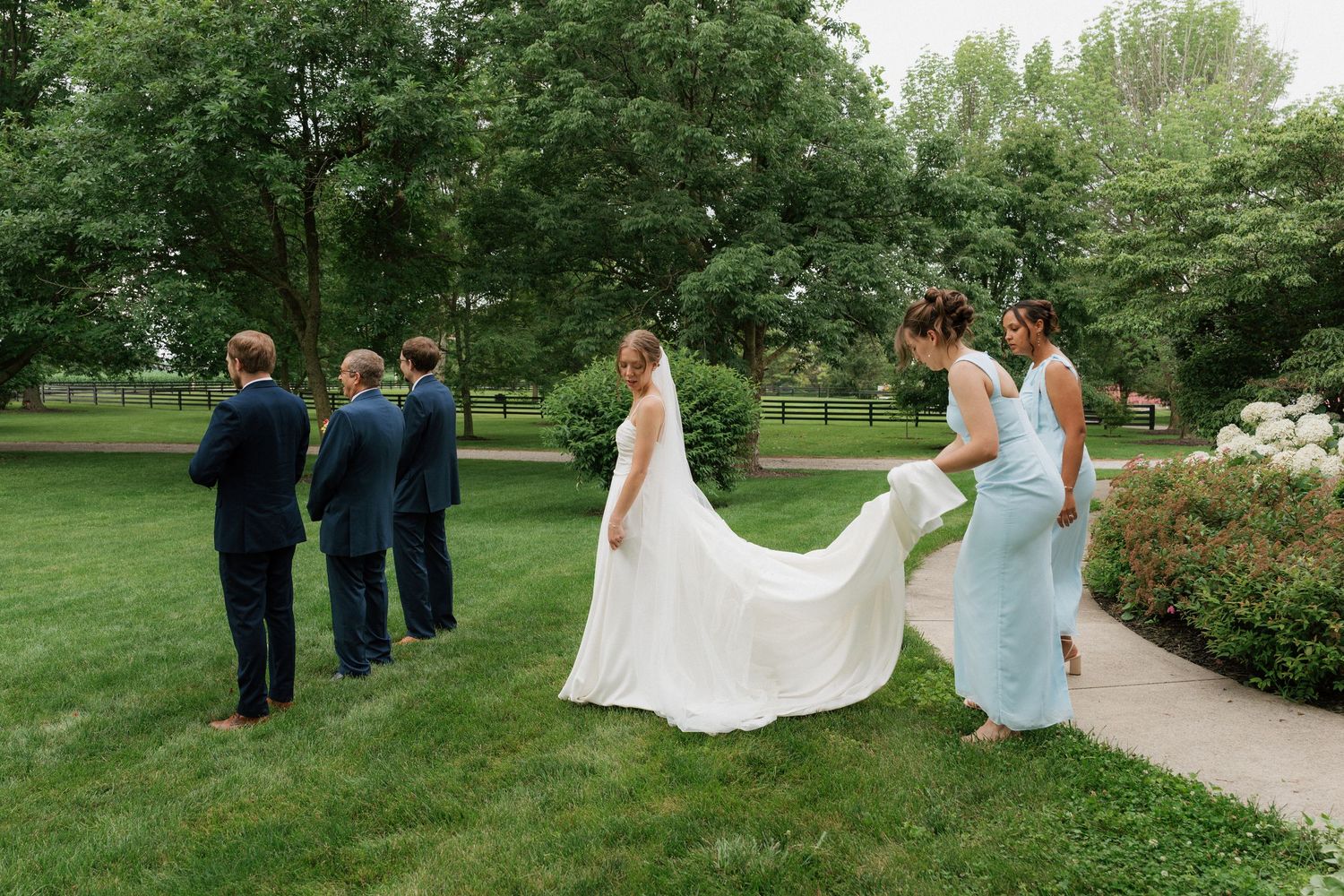 Bride in white gown with bridesmaid holding train in lush garden, groomsmen in navy suits nearby.