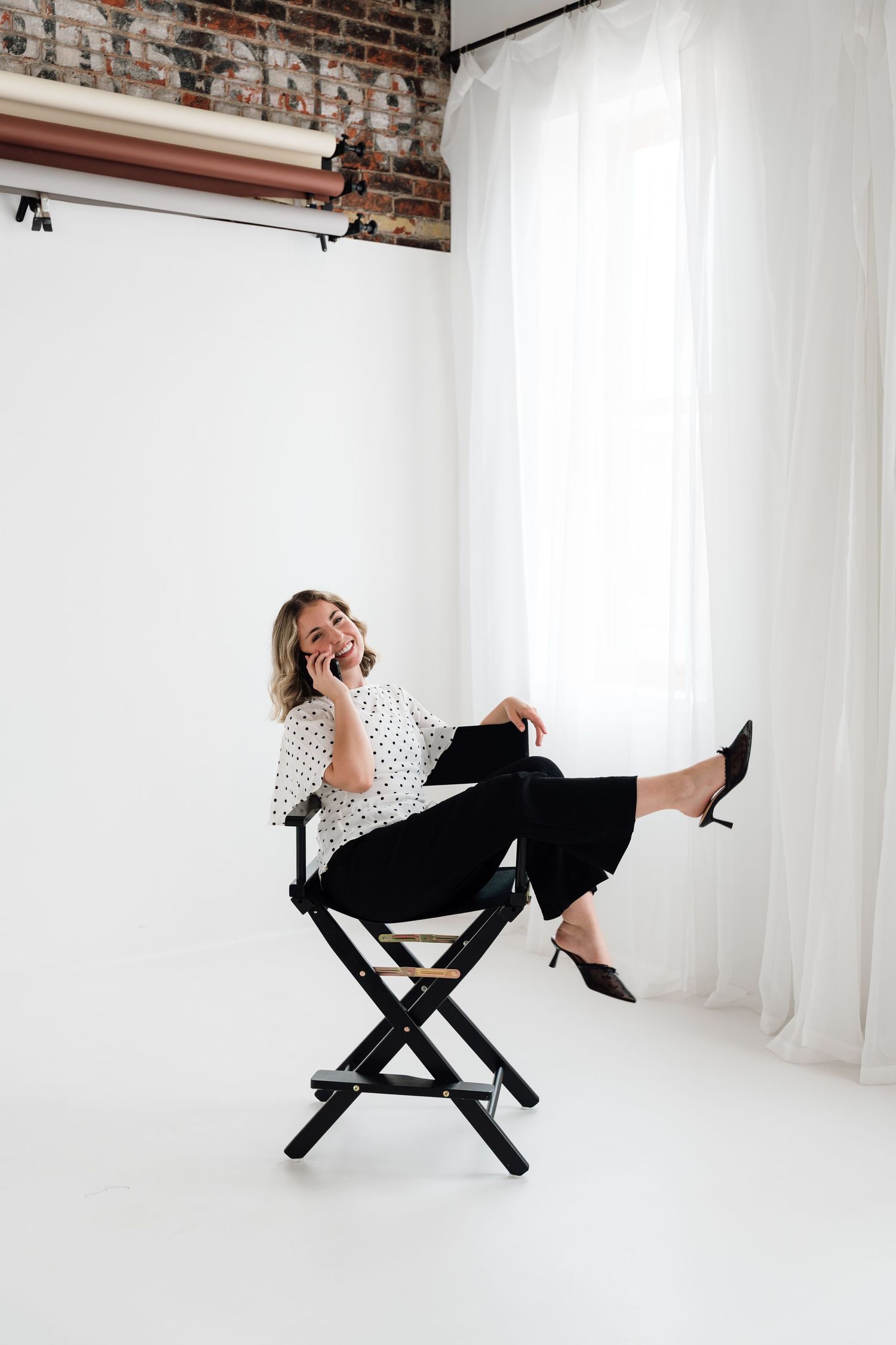 Woman in polka dot blouse and black pants laughing on phone while seated in director's chair in white studio.