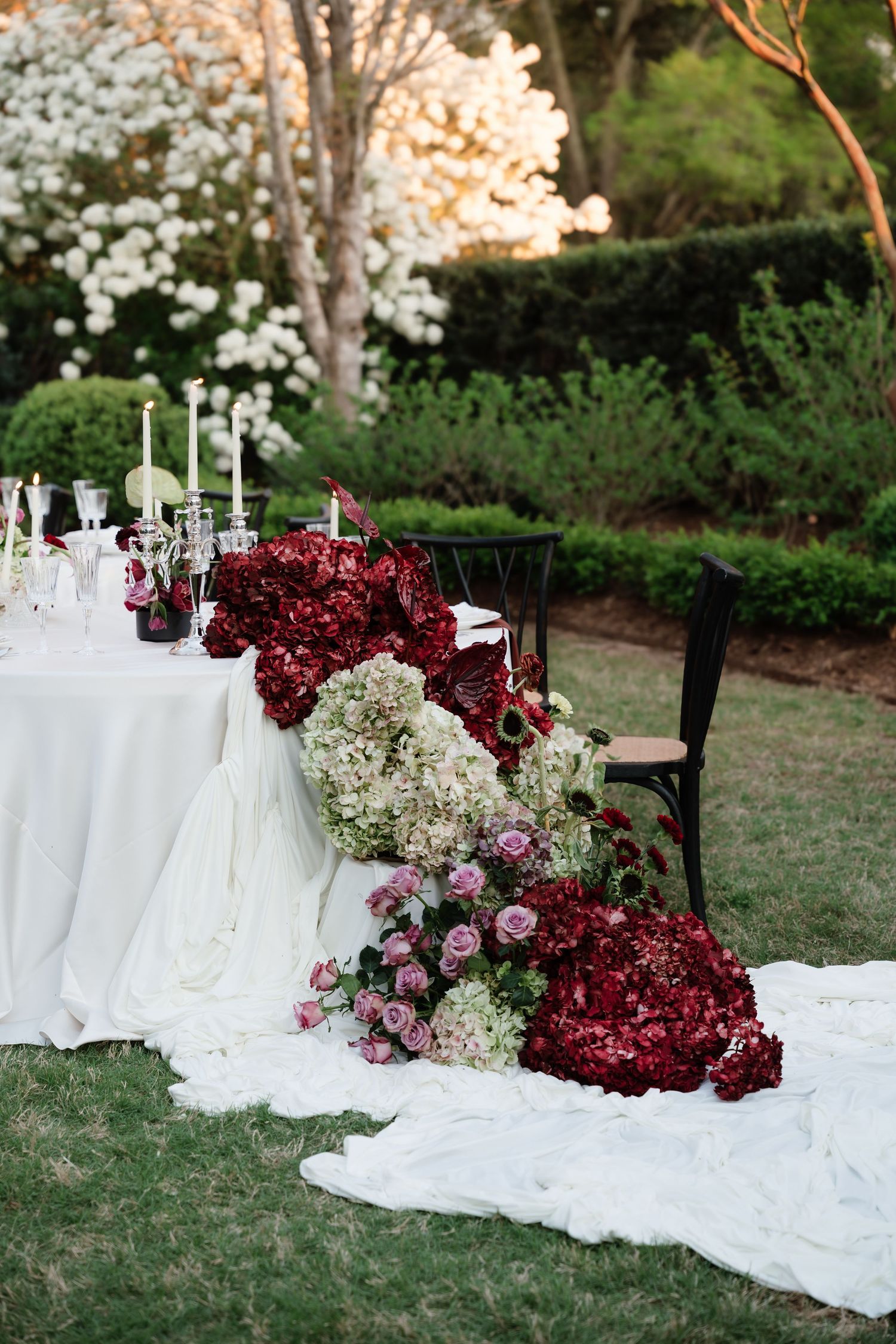 Elegant outdoor wedding tablescape with burgundy hydrangeas, white florals, candles, and cascading blooms on white linen.