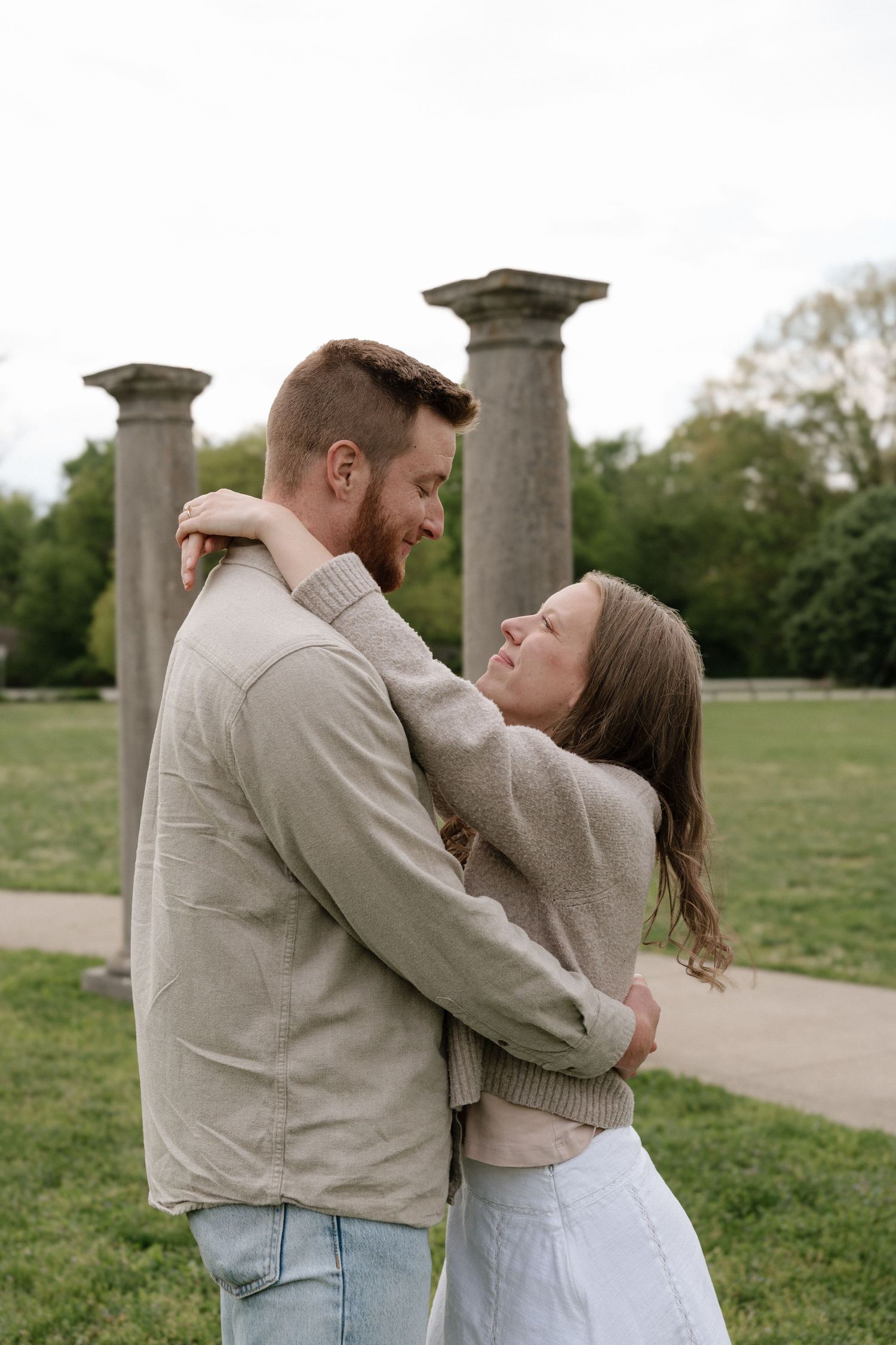 Couple embracing and smiling at each other in a park with stone columns in the background.