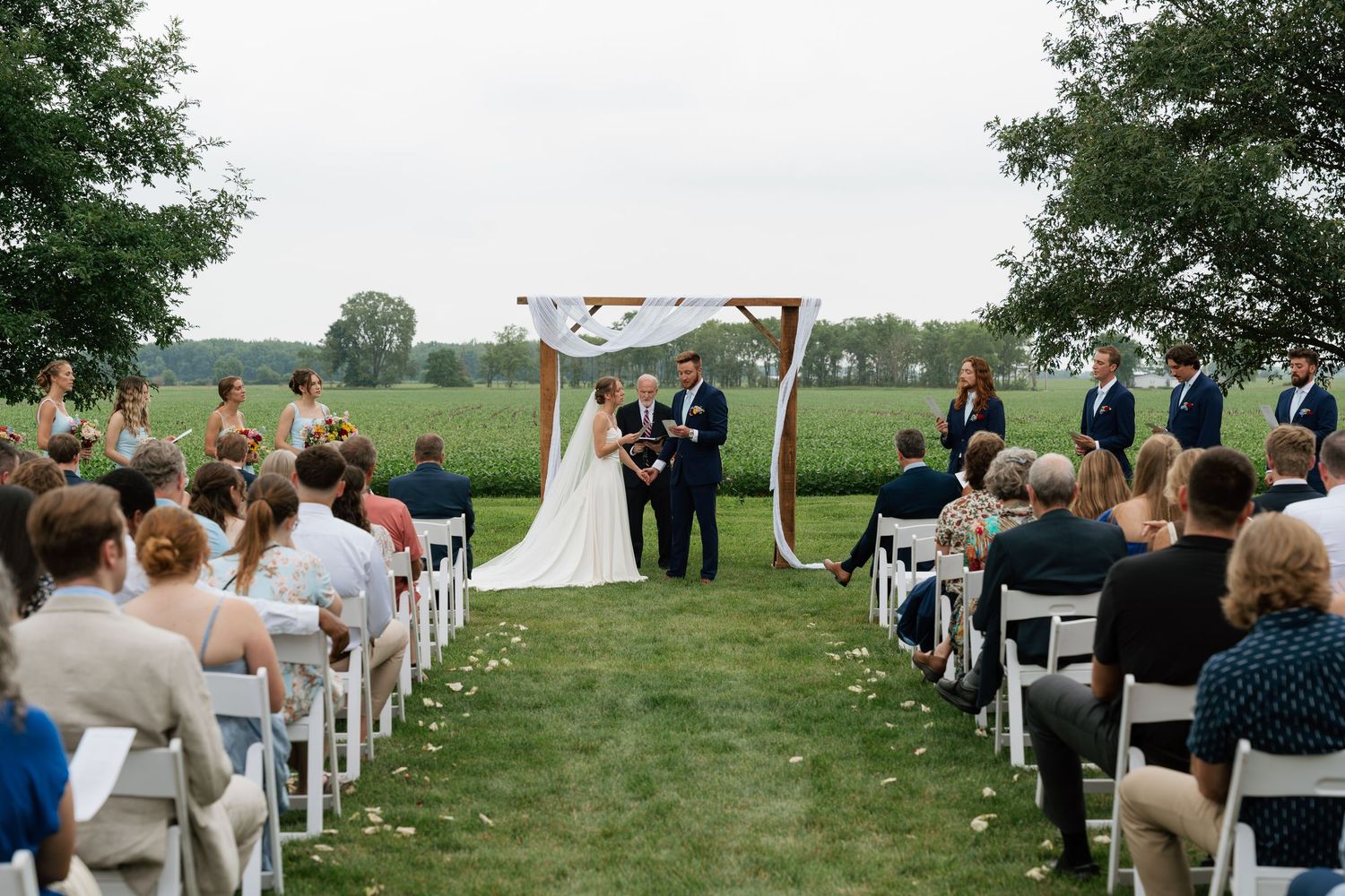 Outdoor wedding ceremony on green lawn with floral arch, bride and groom at altar, guests seated in white chairs.