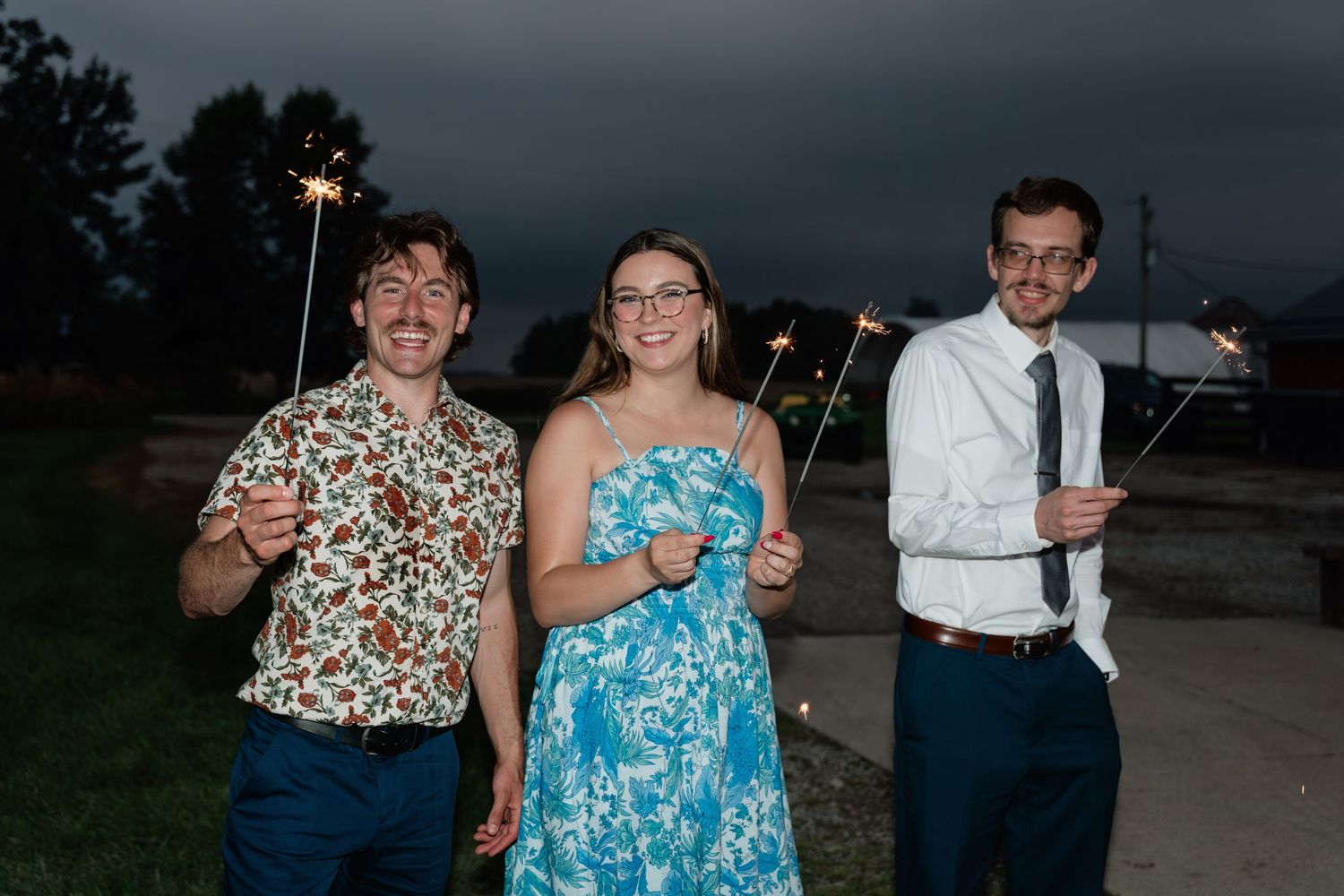 Three friends holding sparklers outdoors at dusk, woman in blue dress flanked by two men smiling at a celebration.
