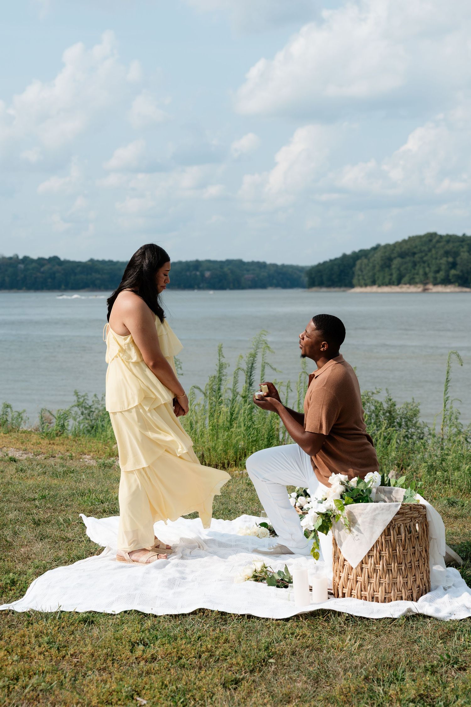 Man proposes to woman on one knee during lakeside picnic, holding ring box with flowers and basket nearby.