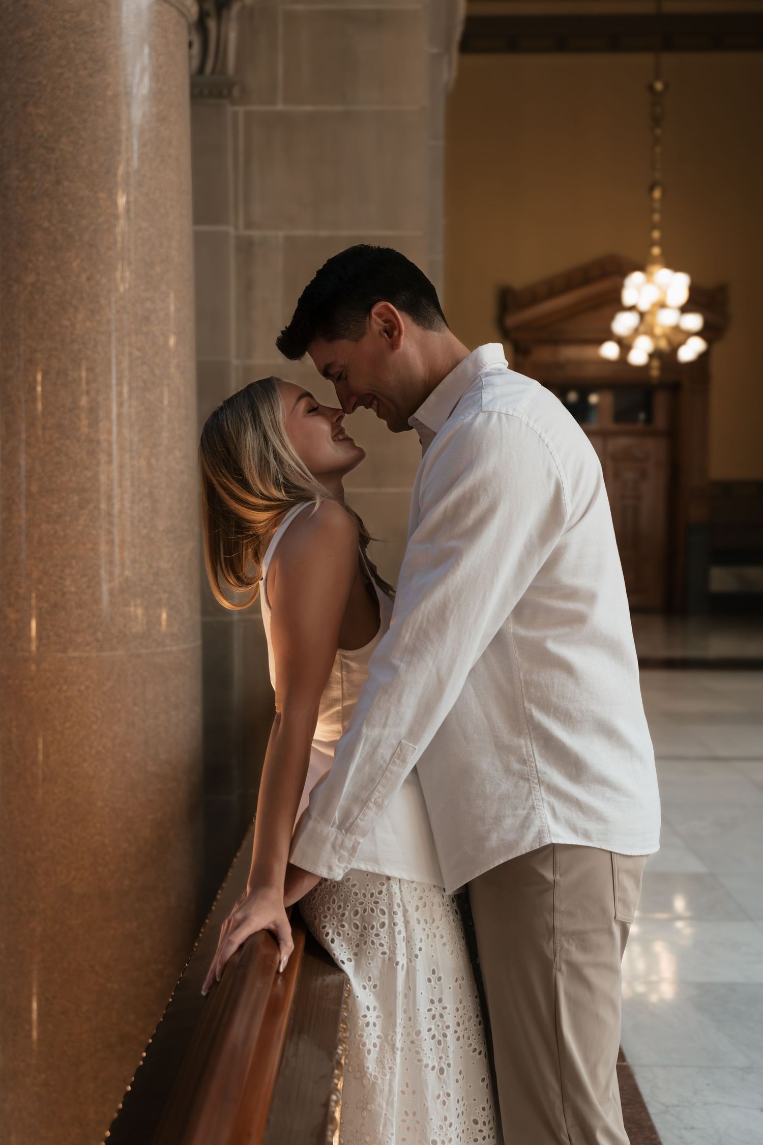 Romantic couple in white outfits sharing an intimate moment leaning against marble columns in an elegant indoor setting.