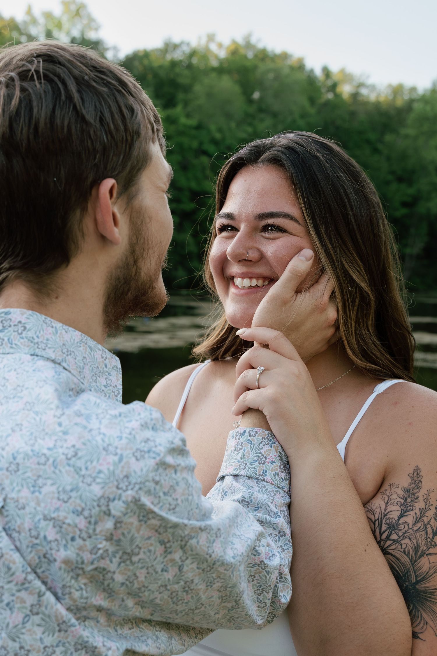 A smiling woman with a tattoo gazes at a bearded man who gently cups her face near a serene lake backdrop.