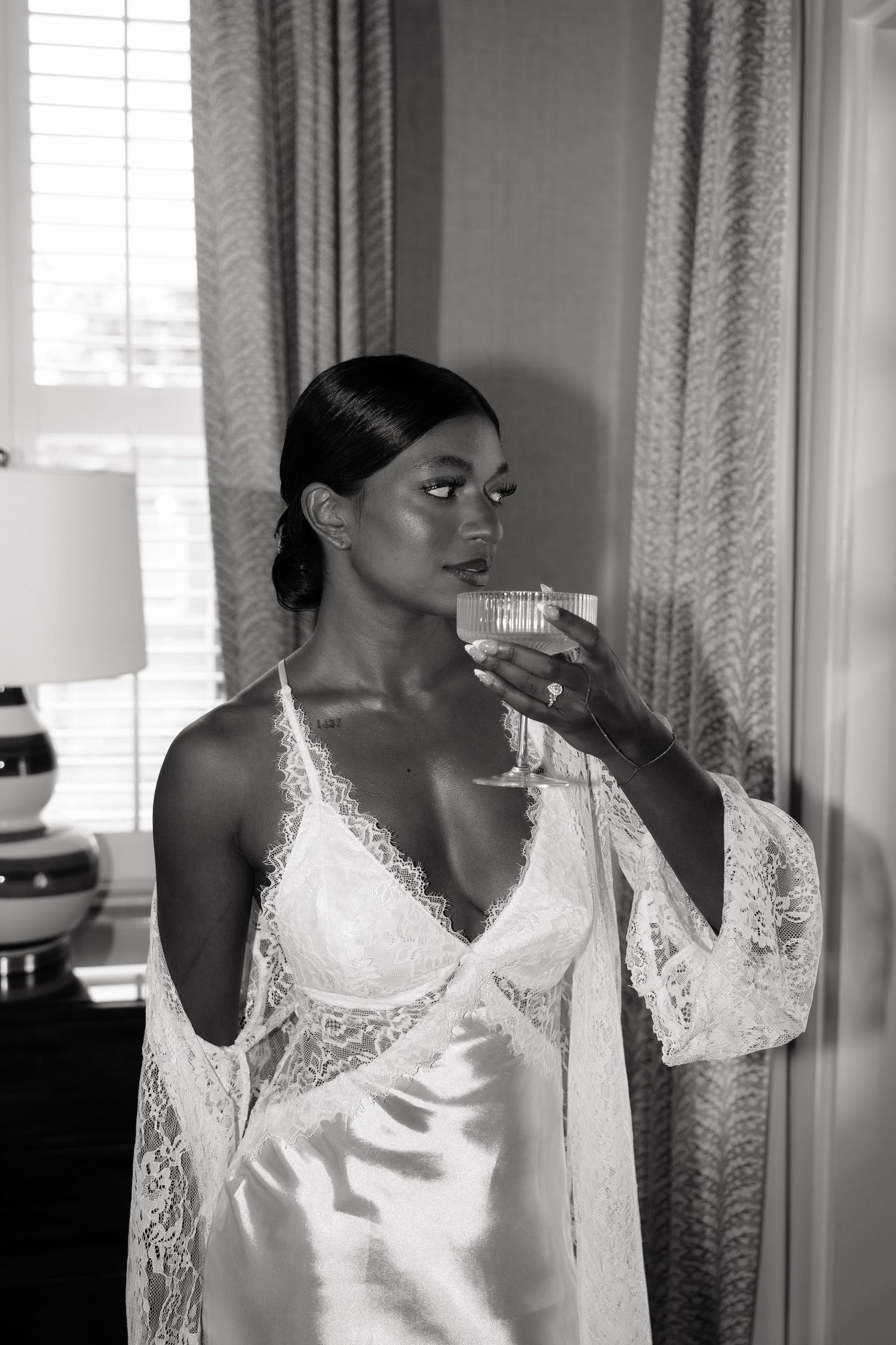 Black and white photo of elegant woman in lace bridal lingerie holding champagne glass near window.