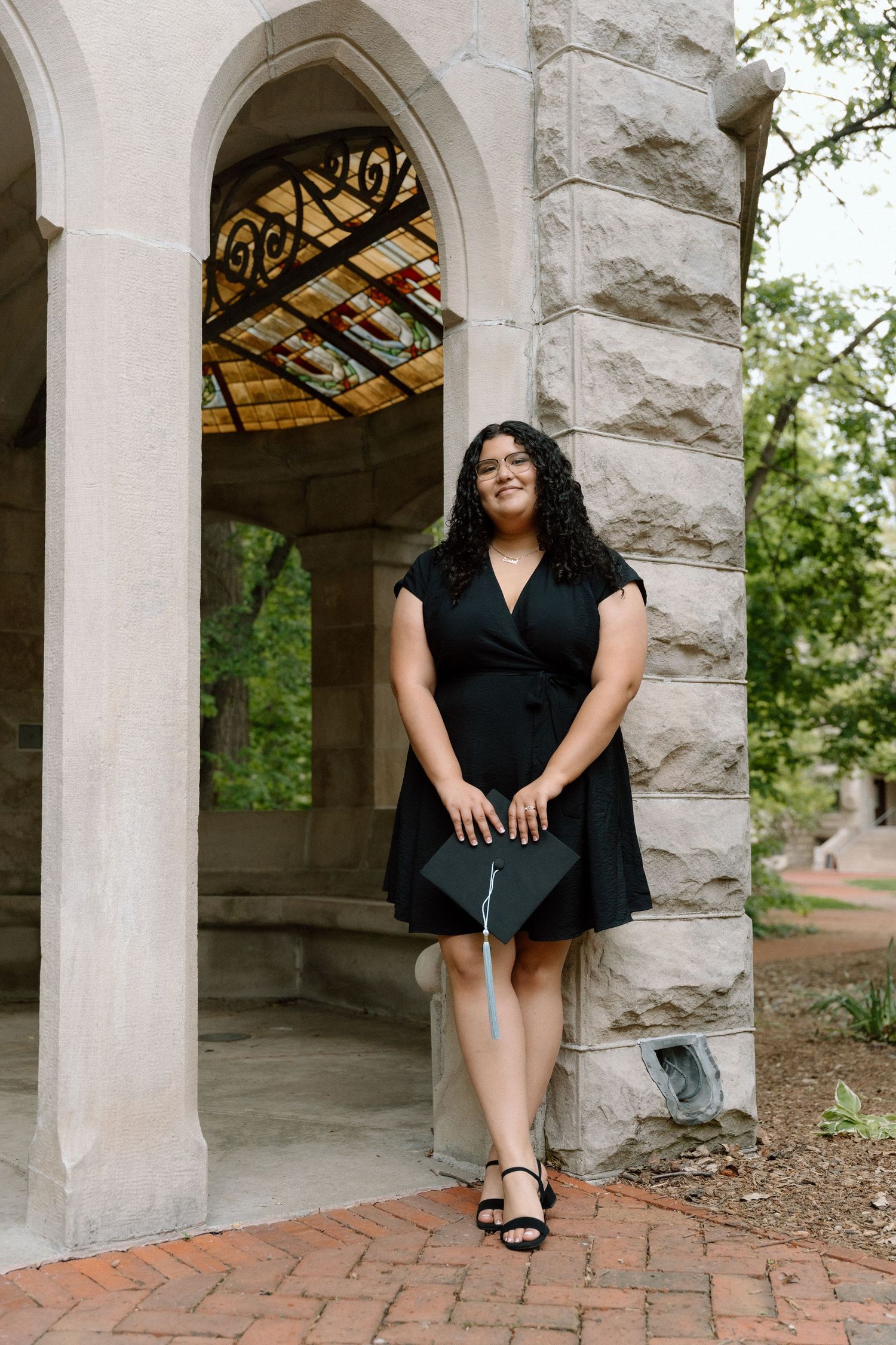 A young woman in a black dress holds a graduation cap while posing by stone arches at a university.