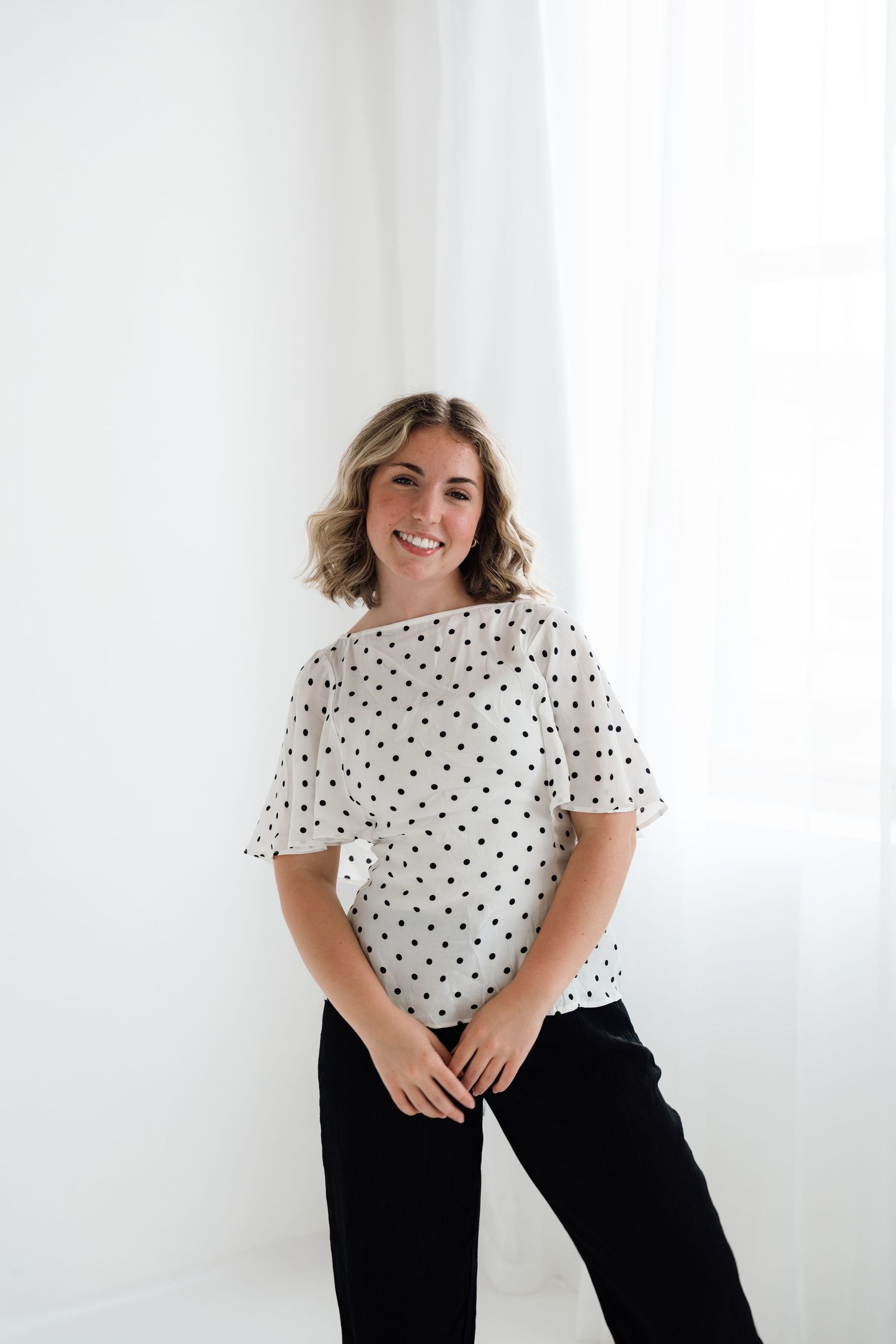Smiling woman wearing white polka dot blouse and black pants standing against white background.