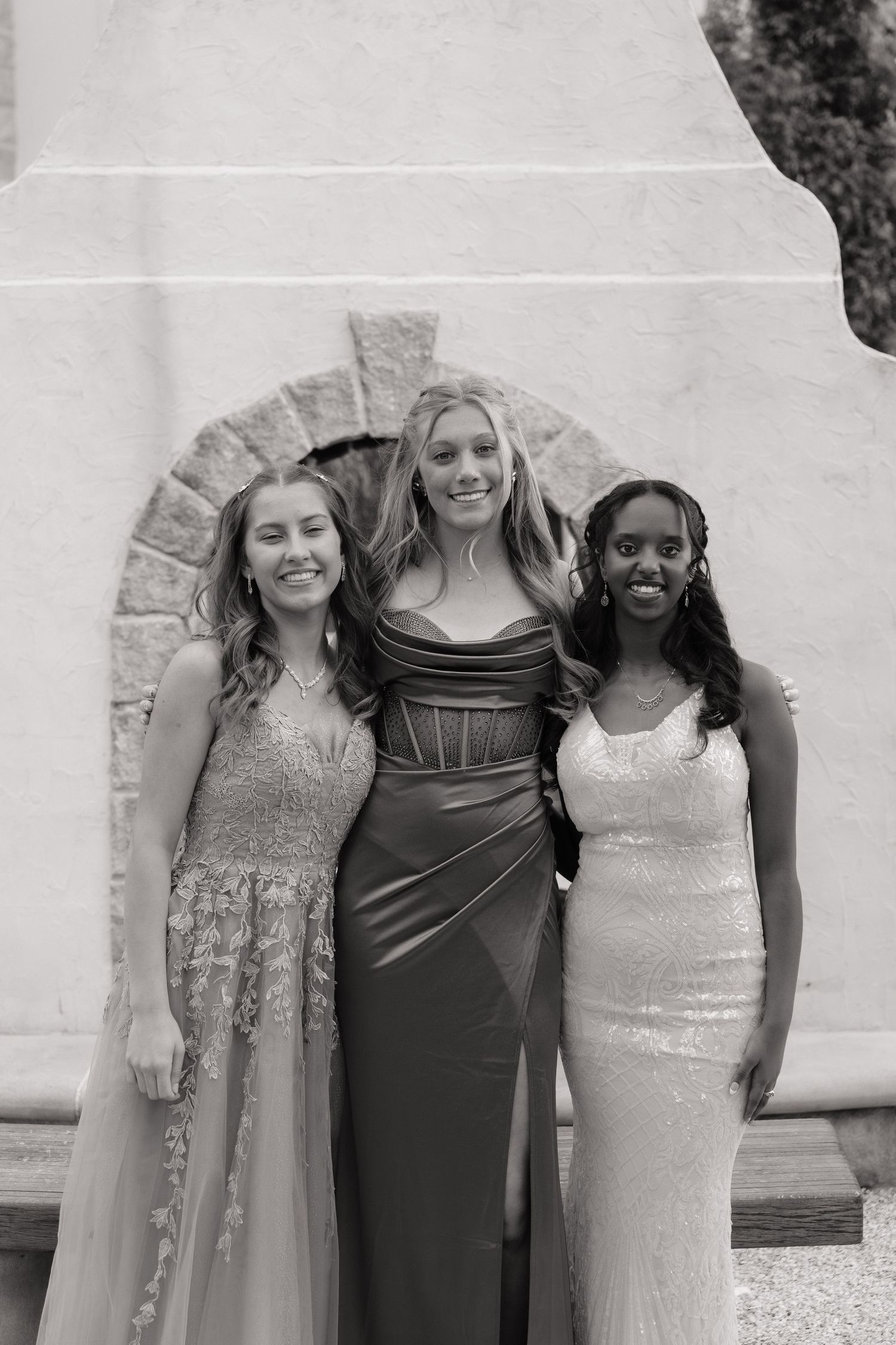 Three young women in elegant formal gowns pose together smiling in a black and white prom photo.