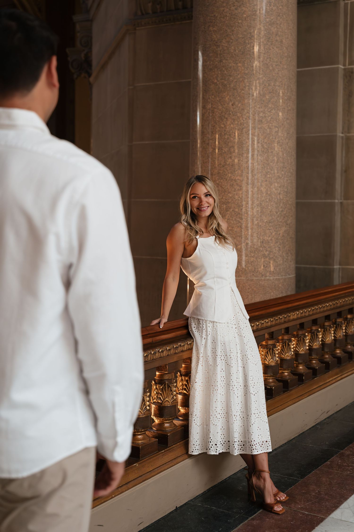 Woman in white lace skirt and top smiles at man in elegant marble hall during engagement photo session.
