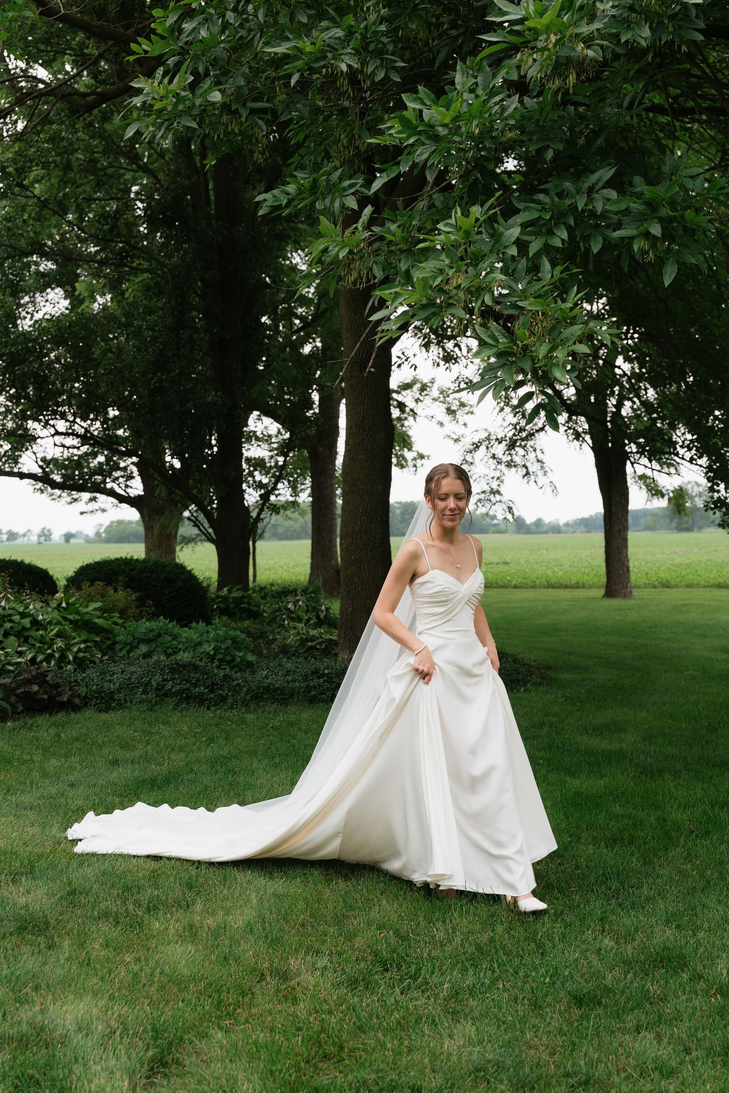 Elegant bride in strapless ball gown with long train poses gracefully under large trees outdoors.