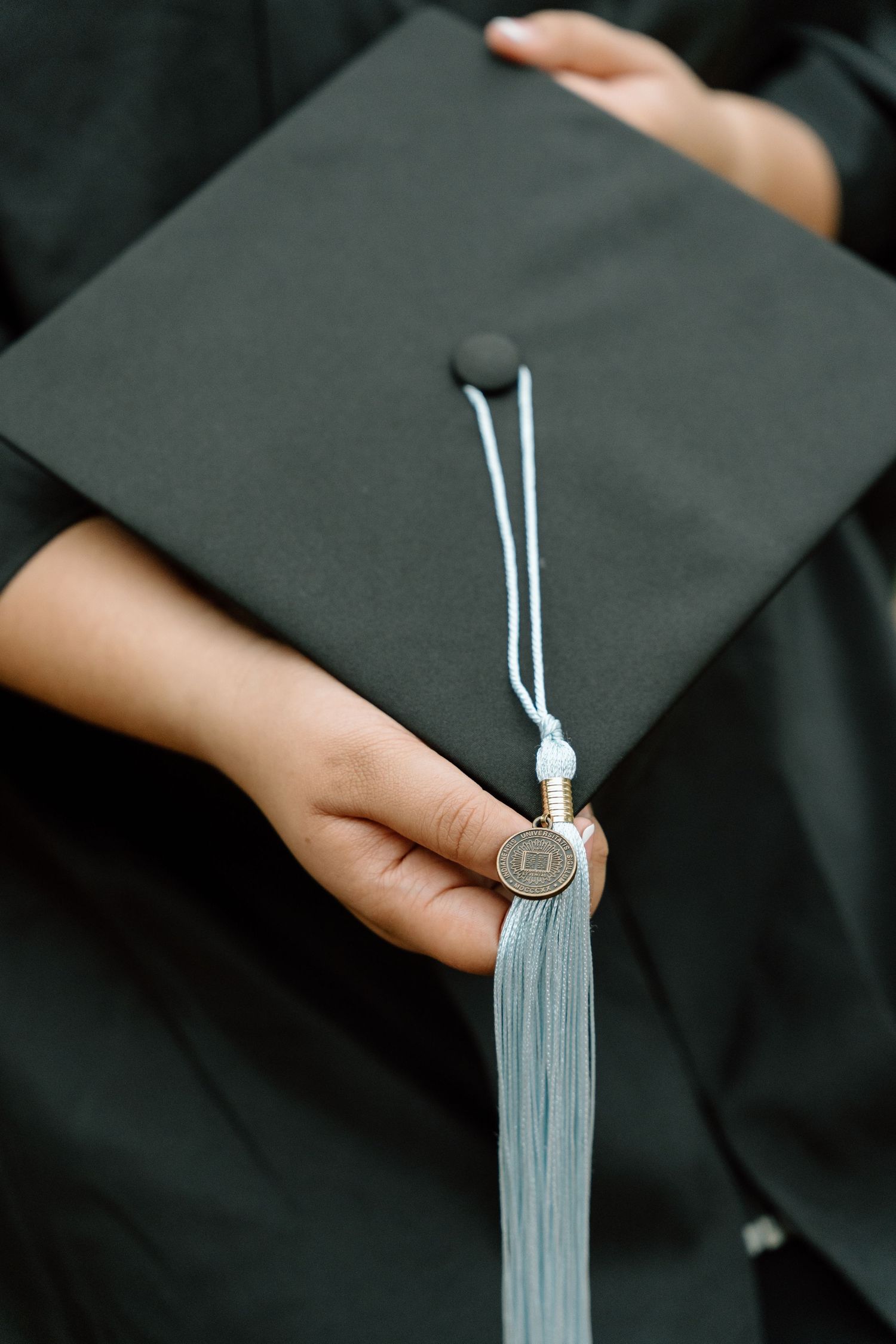 A graduate holds a dark green mortarboard cap with a light blue tassel and gold medallion during graduation ceremony.
