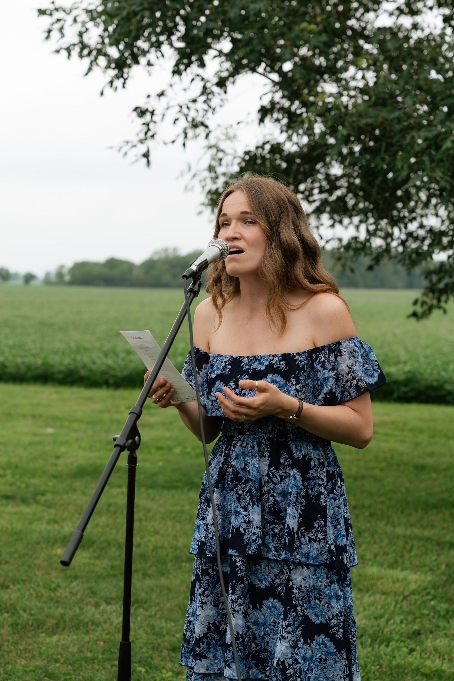 Woman in blue floral off-shoulder dress speaking at outdoor microphone stand holding notes at garden event.