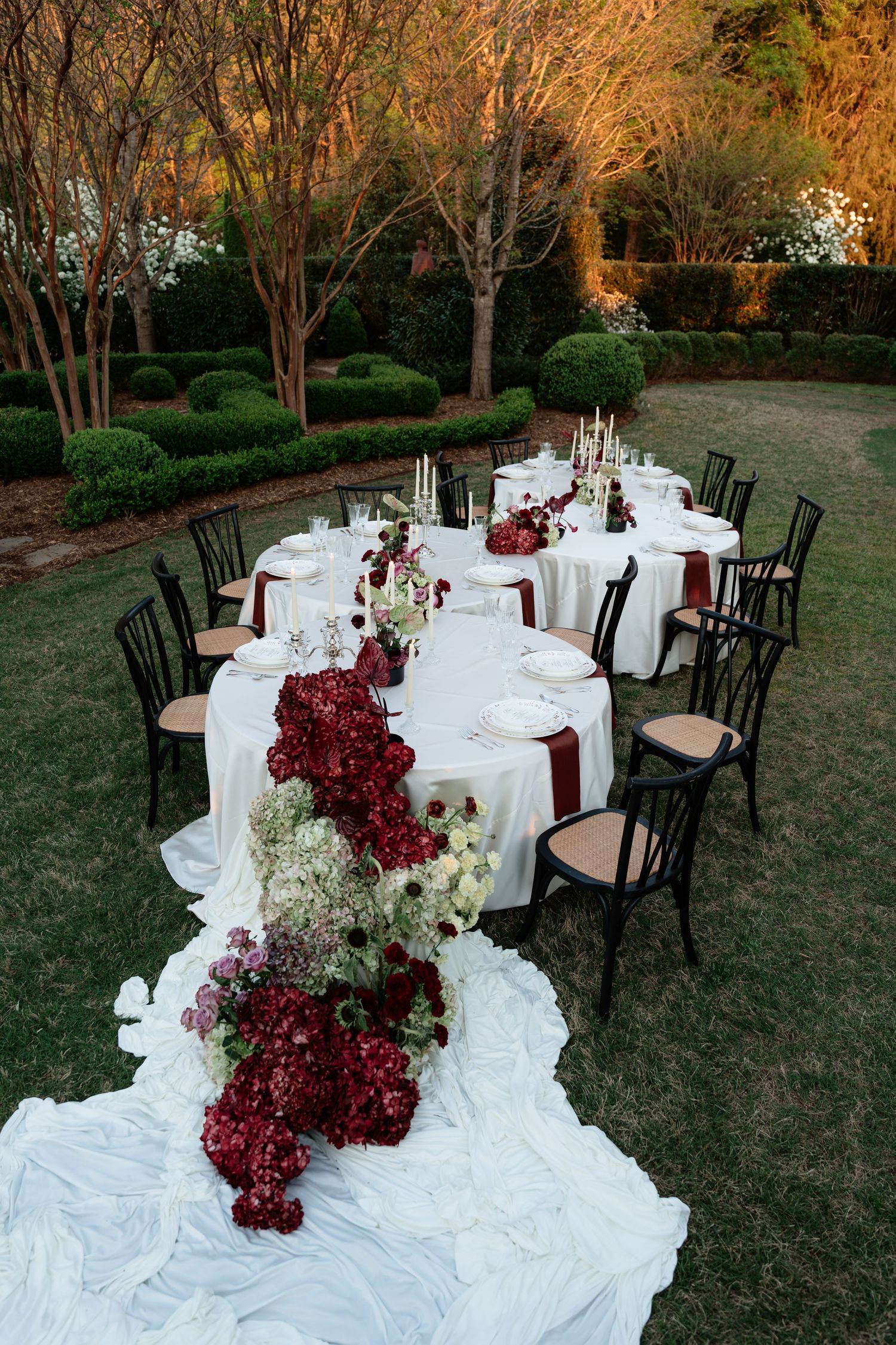 Outdoor wedding reception with white tablecloths, black chairs, and cascading burgundy floral arrangements on lush green lawn.