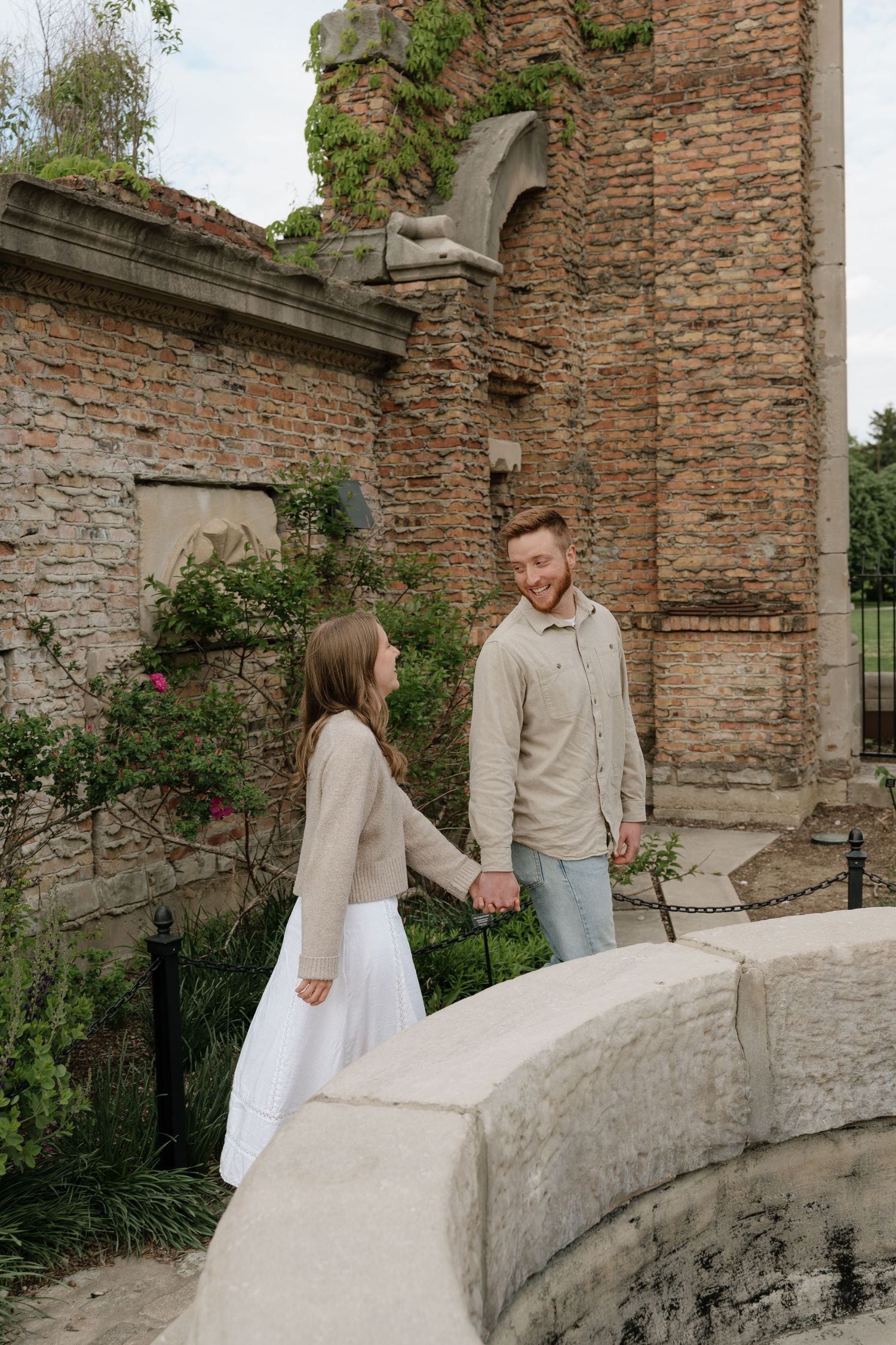 Couple holding hands on stone bridge beside flowering brick ruins in a lush garden.
