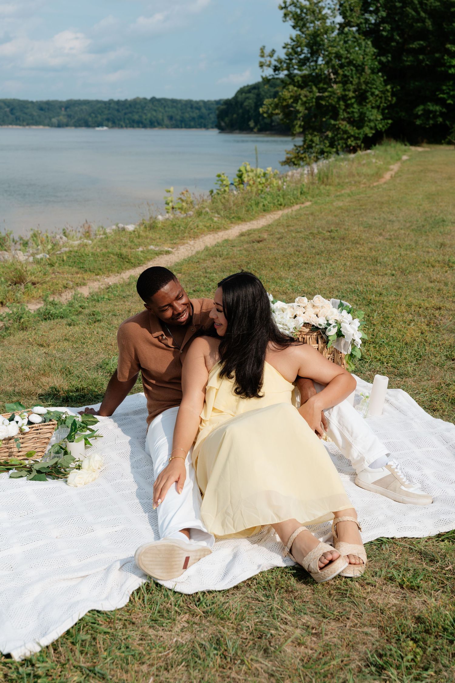 Couple enjoying a romantic lakeside picnic on a white blanket with flowers and a basket on a sunny day.