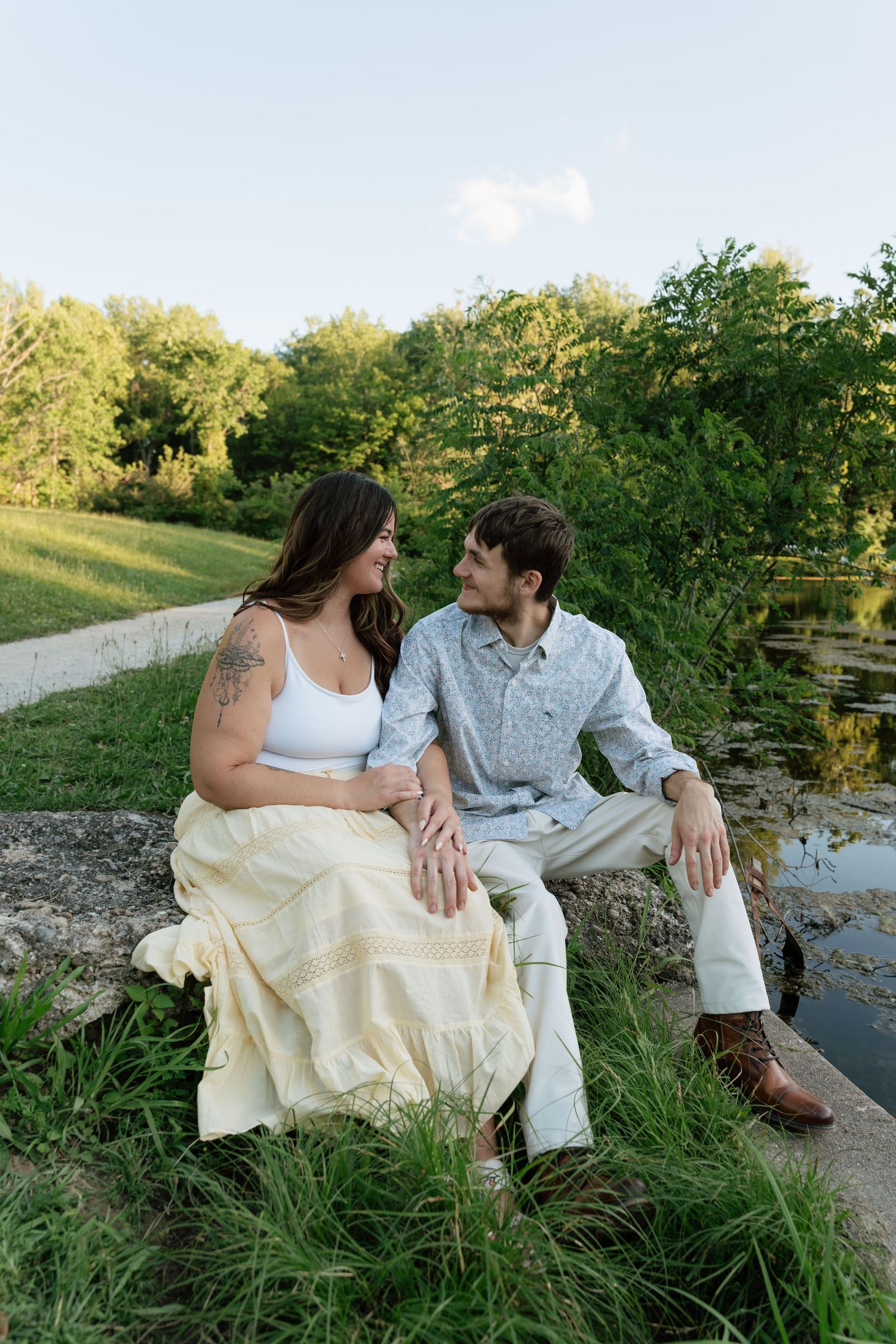 A couple sits together on a stone ledge by a pond, gazing at each other in a lush green park setting.