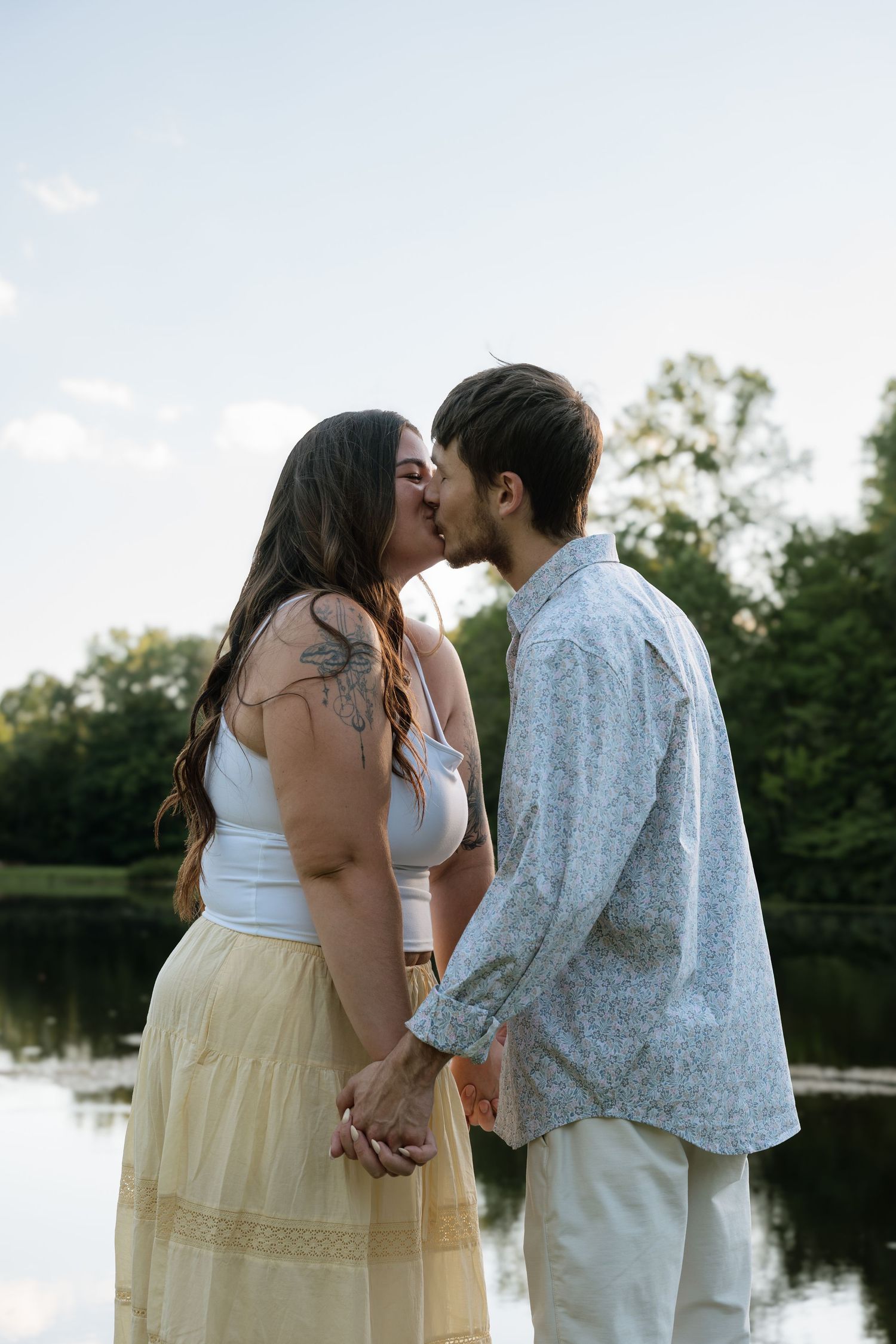 A couple shares a romantic kiss while holding hands near a serene lake surrounded by lush green trees.