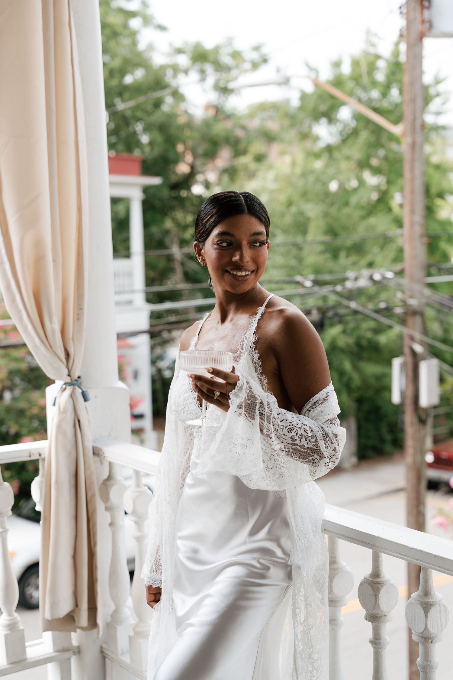 Smiling bride in white satin gown and lace wrap holds champagne glass on elegant white balcony.