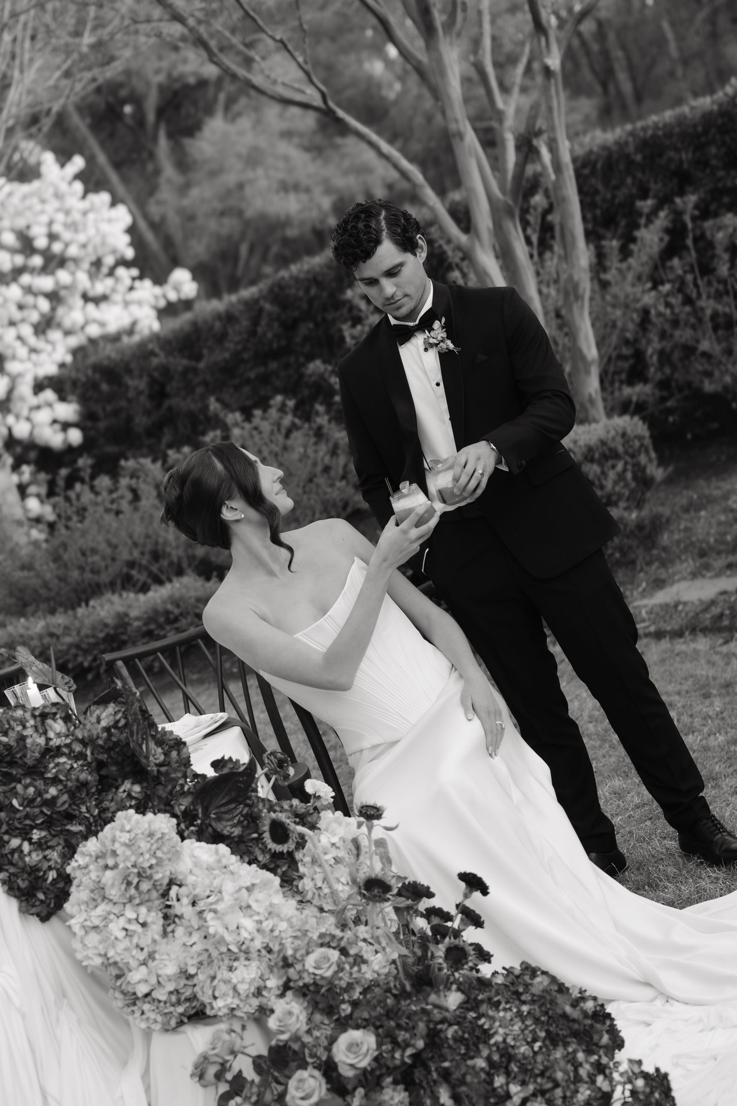 Black and white photo of bride in white gown seated as groom in tuxedo stands beside her near floral arrangements.