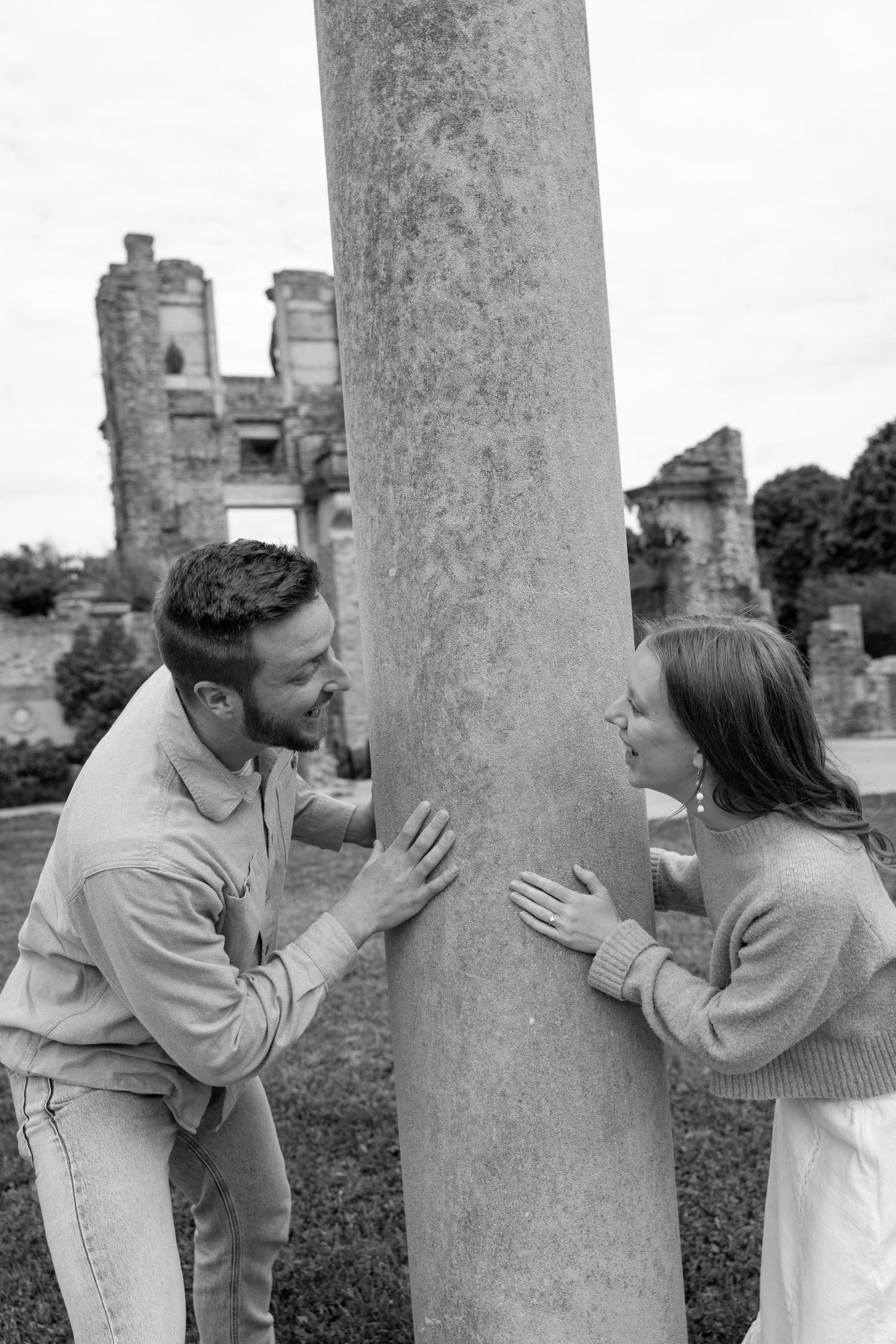 Black and white photo of a couple playfully peeking around a stone column at historic ruins.